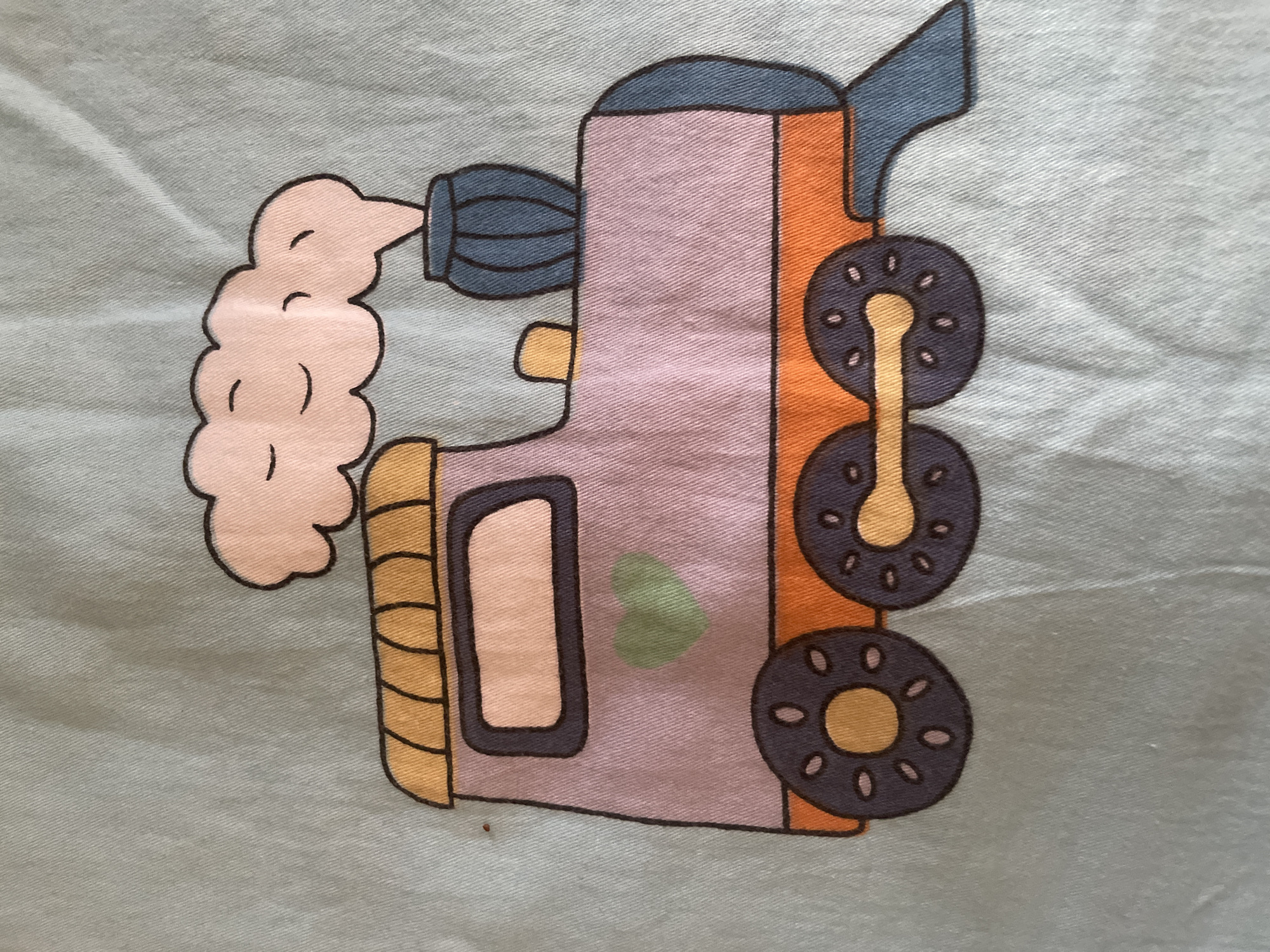The image is a close-up, top-down view of a light blue fabric with a printed illustration of a cartoon train. The train is colored in pastel shades of pink, orange, yellow, and blue, with black outlines. It features a large, pink engine with a yellow roof, a blue smokestack emitting a pink cloud with curved lines representing a face, and large, dark blue wheels with orange centers. A green heart shape is visible on the side of the engine. The fabric shows wrinkles and texture, indicating it is likely clothing or bedding. The scene is devoid of any urban environment or context related to Tashkent, Uzbekistan.