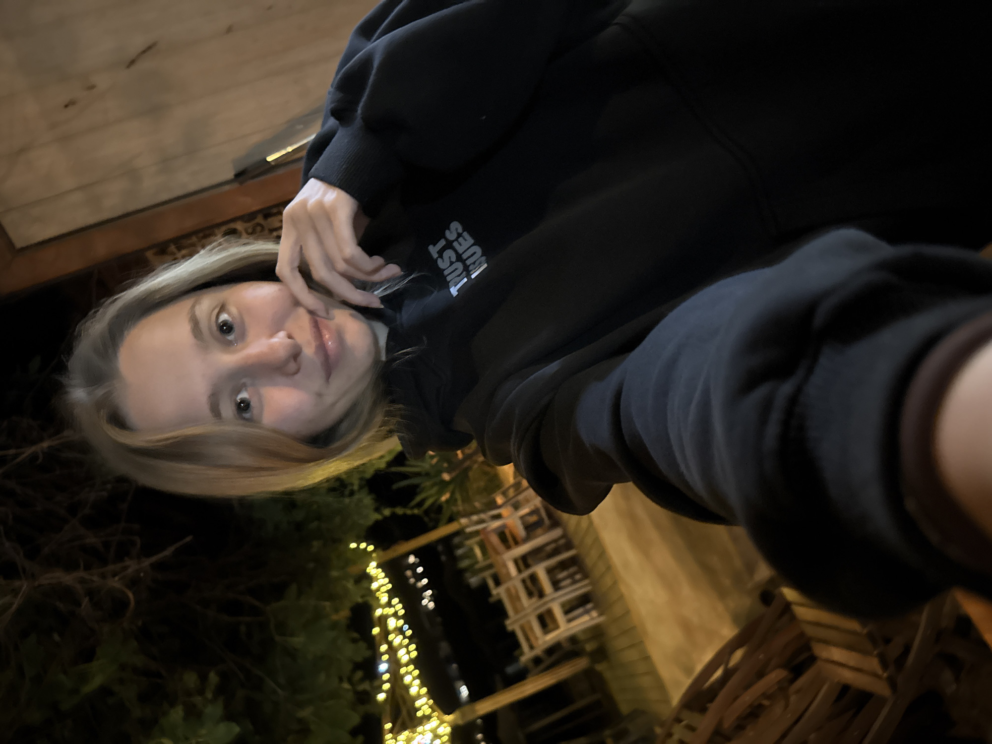 A young blonde woman is taking a selfie at night. She is wearing a black hoodie with the words "Trust Issues" in white lettering across the chest. She is smiling and touching her chin with her left hand. The background is out of focus, but appears to be an outdoor seating area with tables and chairs, likely a restaurant or bar, illuminated by string lights. The overall scene suggests a casual evening outing in Tsarevo, Bulgaria.