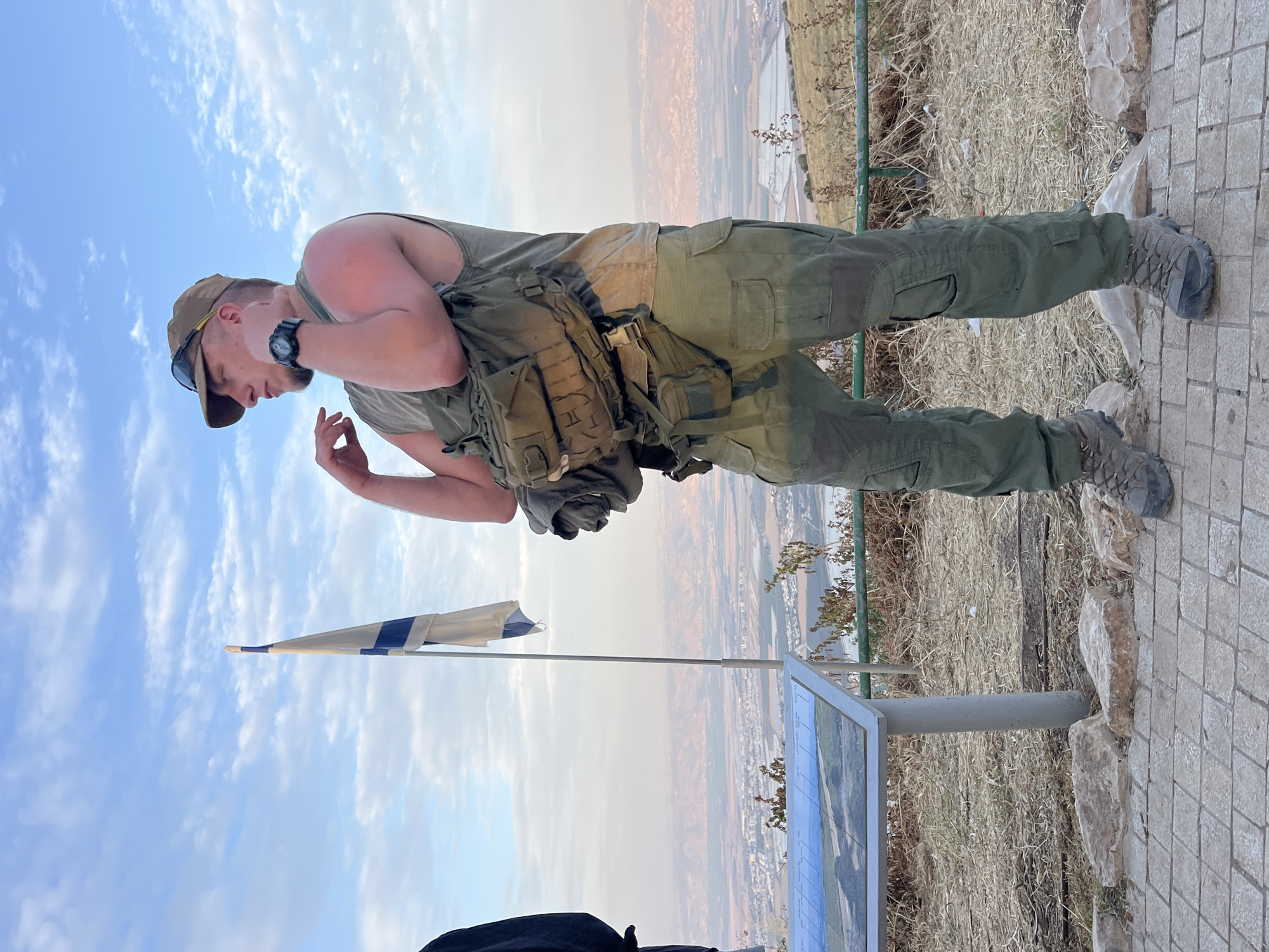 A man wearing a brown baseball cap, sunglasses, green tank top, green pants and boots, stands at a viewpoint in Emek HaMaayanot Regional Council, Israel. He is wearing a tactical vest. He is looking down and gesturing with his left hand. To his left is a metal pole holding a small Israeli flag. Next to the flag pole is a display board with an image. Behind the man is a vista of a city. A metal railing runs along the edge of the viewpoint. The ground is covered in stones and dry grass. The sky is blue with scattered clouds.