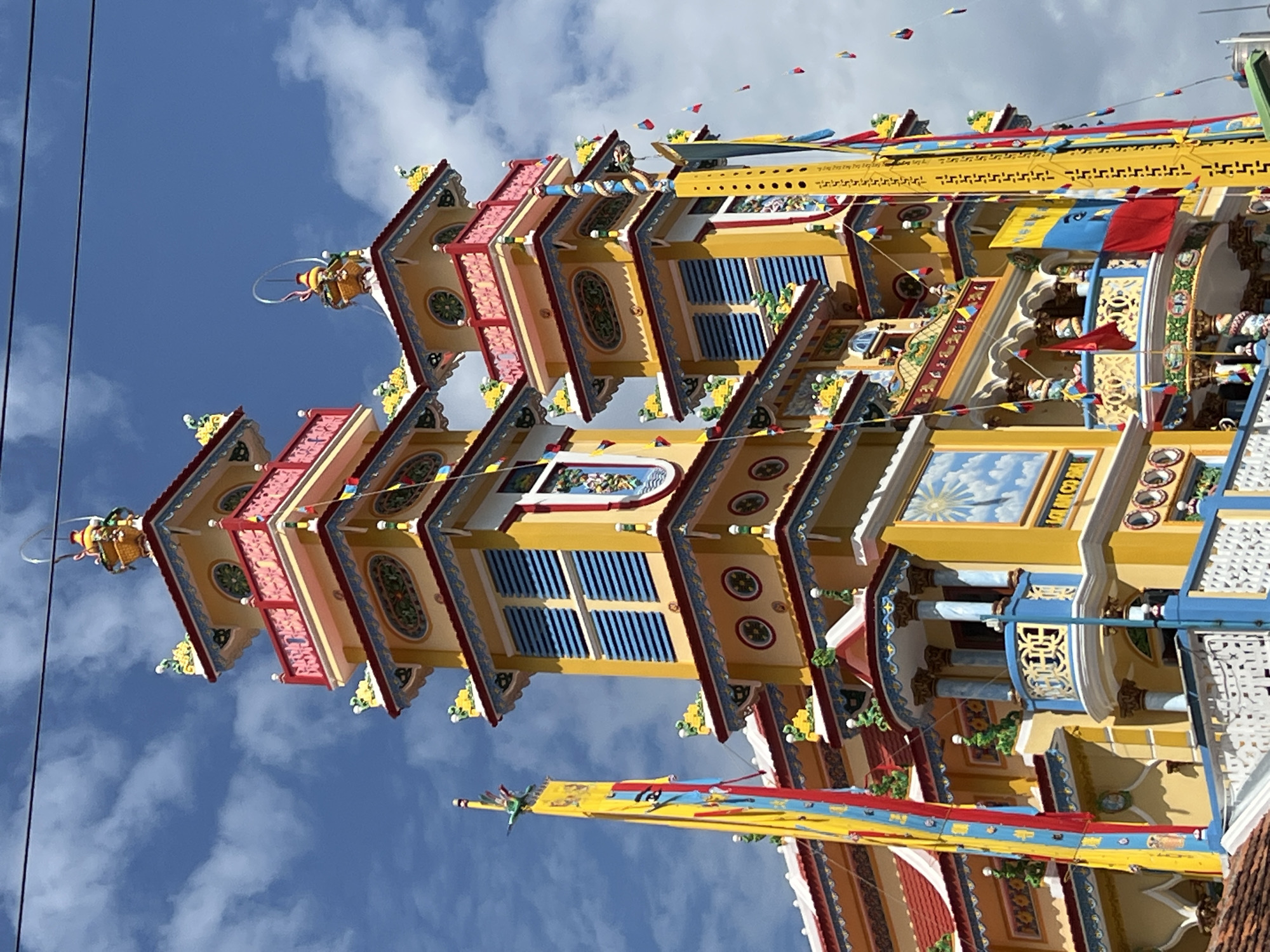 The image shows a close-up, low-angle view of the ornate facade of a colorful religious building in Nha Trang, Vietnam. The building is predominantly yellow with accents of red, blue, and gold. It features multiple tiered roofs, intricate carvings, balconies with decorative railings, and tall, slender towers topped with golden finials. Several long, vertical banners in yellow, blue, and red, along with smaller, triangular flags in similar colors, are prominently displayed on the exterior. The building is set against a bright blue sky with scattered white clouds. The architectural style suggests it is likely a Buddhist temple or a similar religious structure. The overall impression is one of vibrant and elaborate decoration.