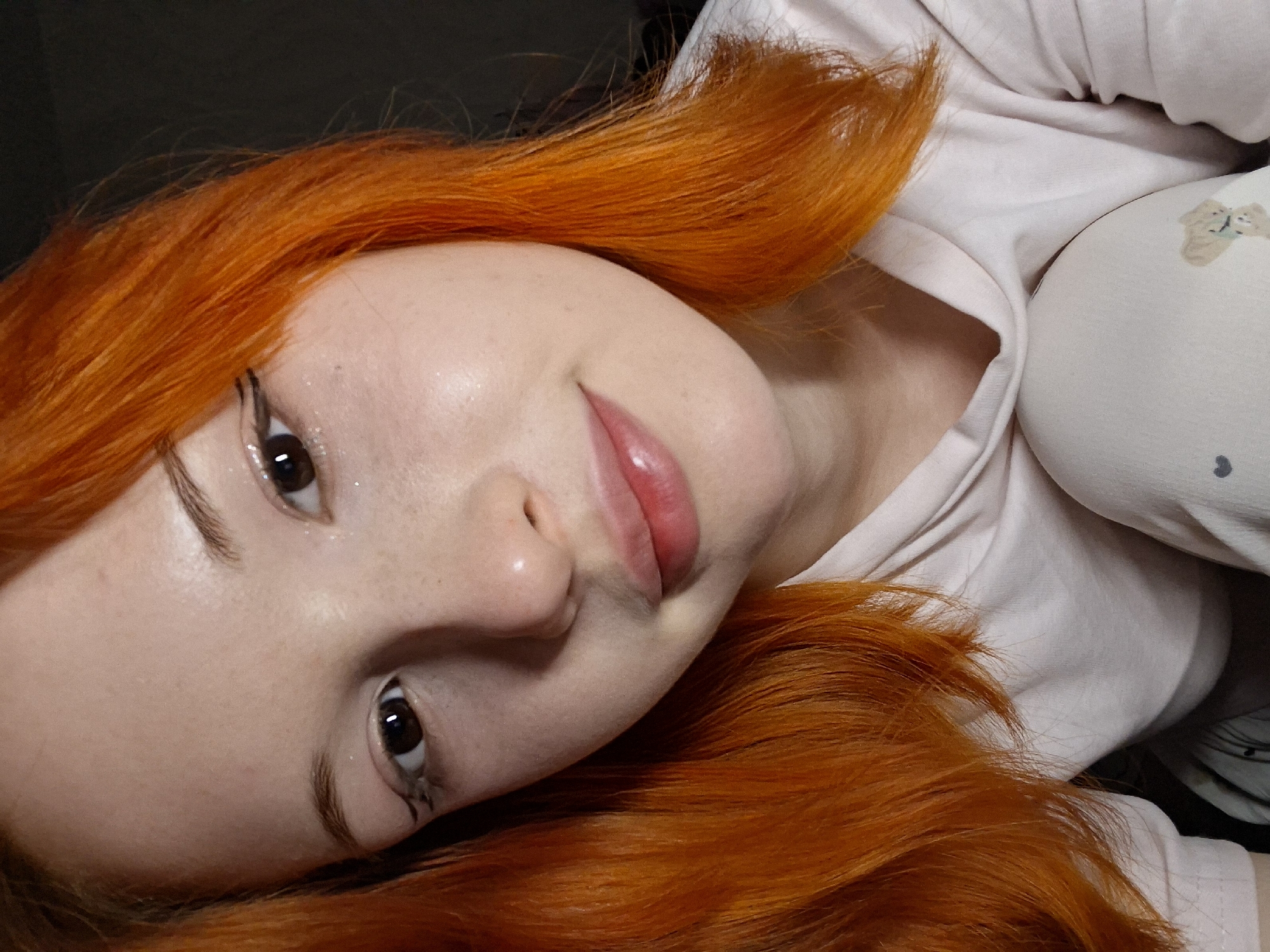 A close-up, angled portrait captures a young female with vibrant, long orange-red hair, likely lying down indoors. Her head is tilted to the left, and she looks directly at the camera with a gentle, soft smile.
The subject has fair skin with subtle freckles. Her dark brown eyes are bright, and hints of sparkle or glitter are visible on her eyelids and below her lower lash lines. Eyeliner, styled with a distinct, slightly artistic wing or line, is present on both upper and lower lids. Her eyebrows are naturally defined. Her lips are a soft rose-pink, slightly parted.
Her striking orange-red hair frames her face, appearing slightly disheveled and fanning out around her head. She is wearing a light-colored, possibly cream or light pink, casual top. To the right side of the frame, a lighter-colored fabric object, likely a pillow or blanket, is partially visible. It features a faint, light brown pattern depicting two small, cuddling figures, possibly animals.
The lighting is soft and even, suggesting an indoor setting and an indeterminate time of day. The background behind her hair on the left is dark and out of focus, while the right side is dominated by the light-colored fabric. The overall mood is calm and serene. There is no visible text in the image. The location context provided is Chaikovsky, Russia.