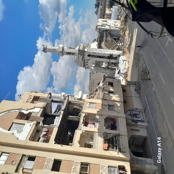 This image depicts a scene of extensive structural damage in Khan Yunis, Palestinian Territory, under a partly cloudy blue sky. On the left, a multi-story, light-toned building stands partially demolished, with multiple floors exhibiting large sections of missing walls, collapsed balconies, and exposed interiors. Laundry is visible hanging from several damaged balconies. Across a debris-strewn paved street, a mosque structure, primarily white, is extensively damaged; its minaret remains partially standing but shows significant structural compromise, while the main body of the mosque is largely reduced to rubble. Piles of concrete and other construction debris are scattered across the street and around the damaged buildings. Two individuals are visible on the street: one figure is located on the left side of the road near a pile of rubble, and a smaller figure walks further down the street in the distance.
