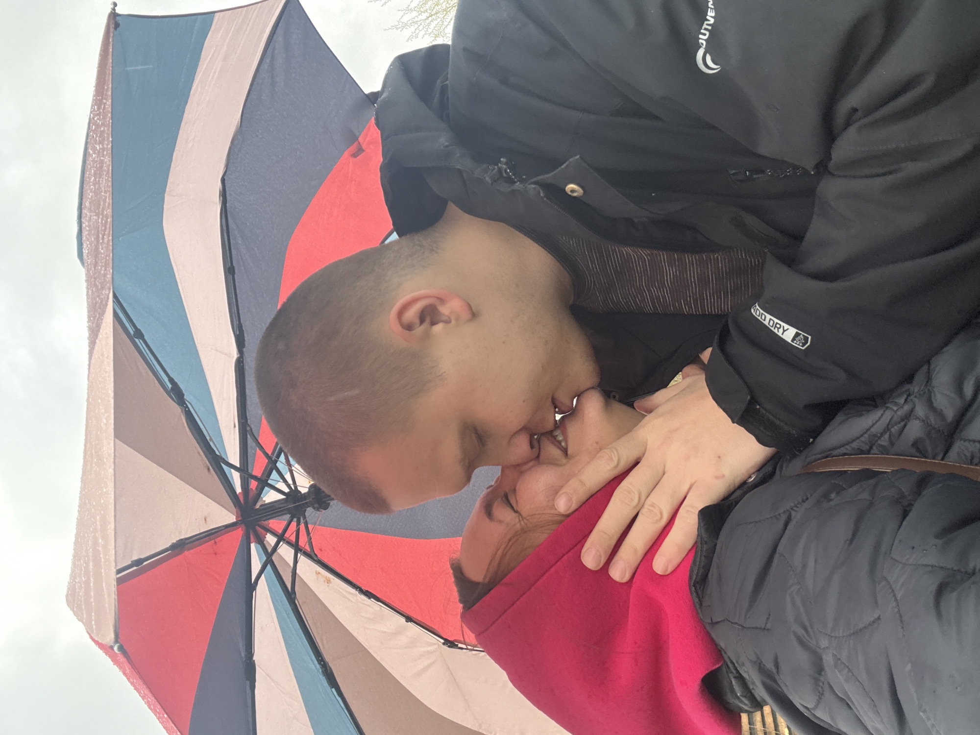 This image captures a man and a woman sharing an intimate kiss under a colorful umbrella. The shot is taken from a low angle, looking upwards towards the couple and the sky. The man, positioned on the left, has short, buzz-cut hair and is wearing a black weather-resistant jacket over a striped shirt. Visible text on his jacket includes "OUTVEN" with a crescent moon logo near the shoulder, and "GOO DRY" with a triangular symbol on the sleeve. The woman, on the right, is wearing a vibrant red hooded top and a black quilted jacket. Her eyes are closed, and she appears to be smiling during the kiss. The man's right hand is gently placed on her face/neck. Above them, a large, open umbrella provides shelter, its surface dotted with what appear to be raindrops, indicating wet weather. The umbrella features wide panels of red, blue, and a light beige or white. The background shows a bright, overcast sky. The scene captures a tender moment of affection, likely during rain in Dmitrov, Russia.