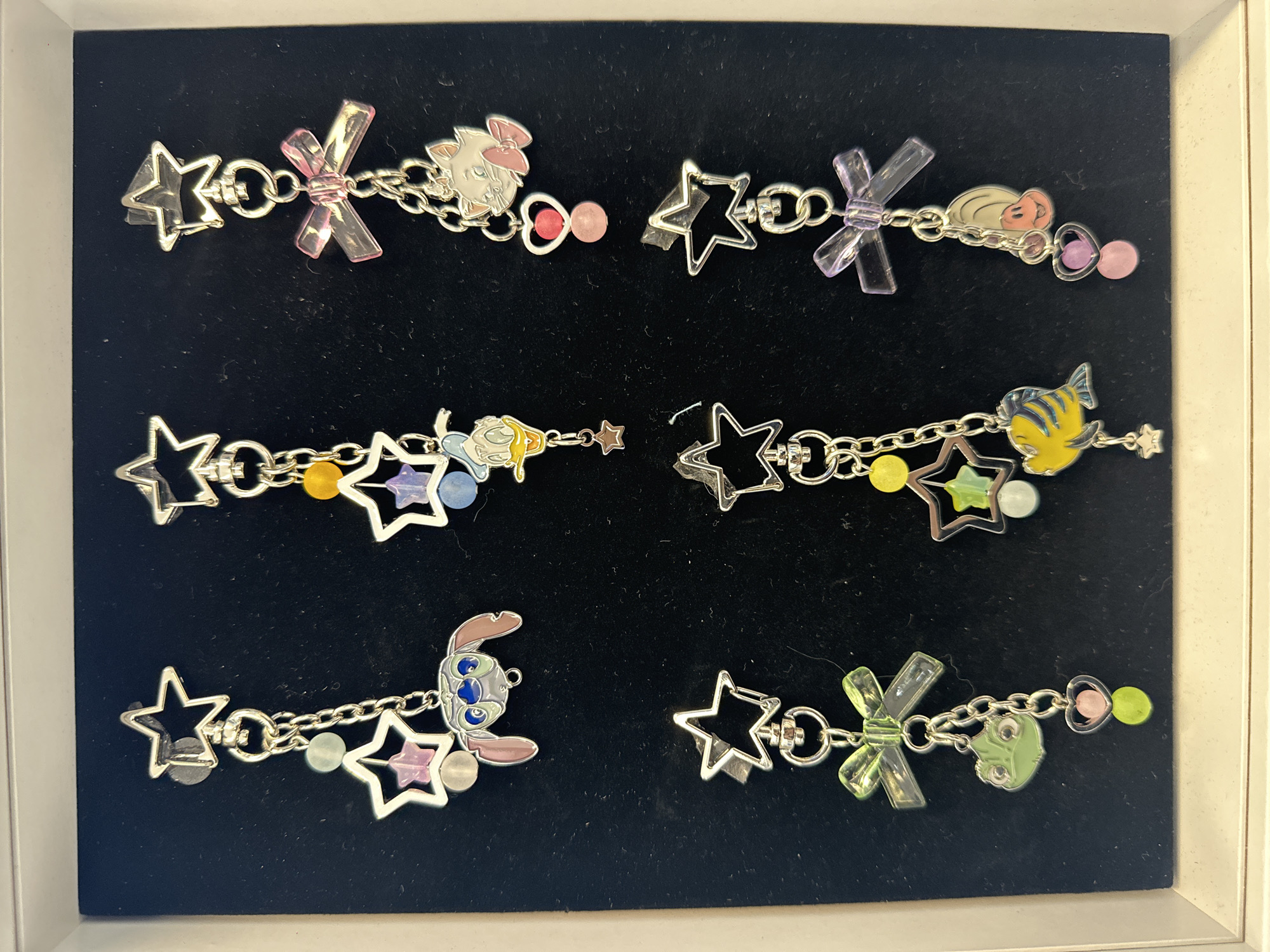 Six Disney-themed charms are displayed vertically on a black velvet background within a white frame. Each charm features a silver-tone star at the top, connected by chains to various Disney characters and embellishments.
From left to right, top row:
1. A charm with Stitch, accompanied by pink and light blue star-shaped beads and a light blue round bead.
2. A charm with Donald Duck, adorned with yellow and purple star-shaped beads and a light blue round bead.
3. A charm featuring Marie from The Aristocats, with a pink ribbon, a heart-shaped charm, and pink and red round beads.
From left to right, bottom row:
4. A charm depicting Kermit the Frog, with a green ribbon and green and pink round beads, along with a heart charm.
5. A charm with Flounder from The Little Mermaid, decorated with light green and light blue star-shaped beads, and a small silver star.
6. A charm with Ariel from The Little Mermaid, featuring a purple ribbon, a heart charm, and pink and purple round beads.
The image appears to be a product display, possibly from a retail store, with no discernible city or specific location details present in the image itself. The items are clean and well-lit.