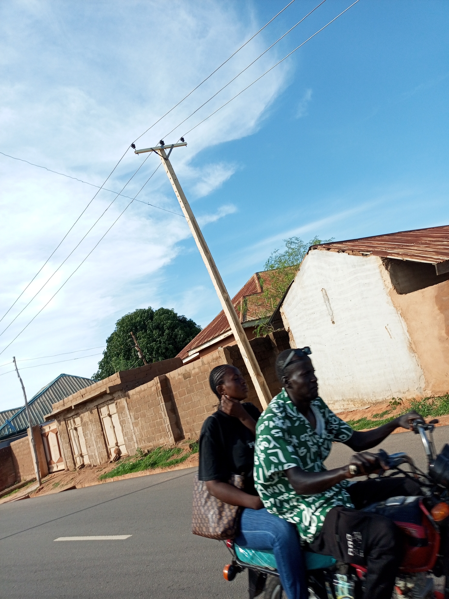 The image shows two people riding a motorcycle in Gombe, Nigeria. The driver, a man wearing a green and white patterned shirt and black pants, is on the right. The passenger, a woman wearing a black shirt and blue jeans, is seated behind him. They are on a paved road with a white line marking a lane. In the background, there are buildings made of brick with corrugated metal roofs, and a large green tree. Telephone poles with wires are visible against a blue sky with scattered white clouds. The scene appears to be in a residential or semi-urban area.
