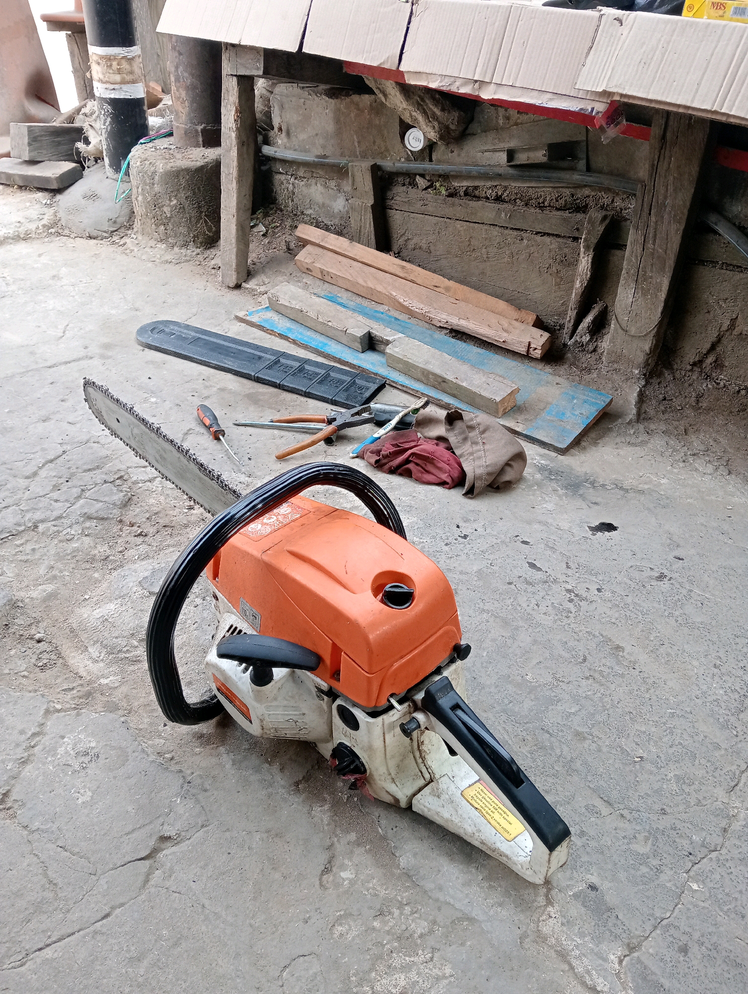 A chainsaw and its guide bar lie on a concrete surface in an outdoor, workshop-like setting. The chainsaw is orange and white with a black handle. The guide bar is black. Several tools, including a screwdriver and pliers, are scattered around the chainsaw. In the background, there are wooden planks, cardboard boxes, and concrete structures, suggesting a work area or storage space. The scene appears to be in Loa Janan Ulu, Indonesia, as indicated by the location. There are no people visible in the image.