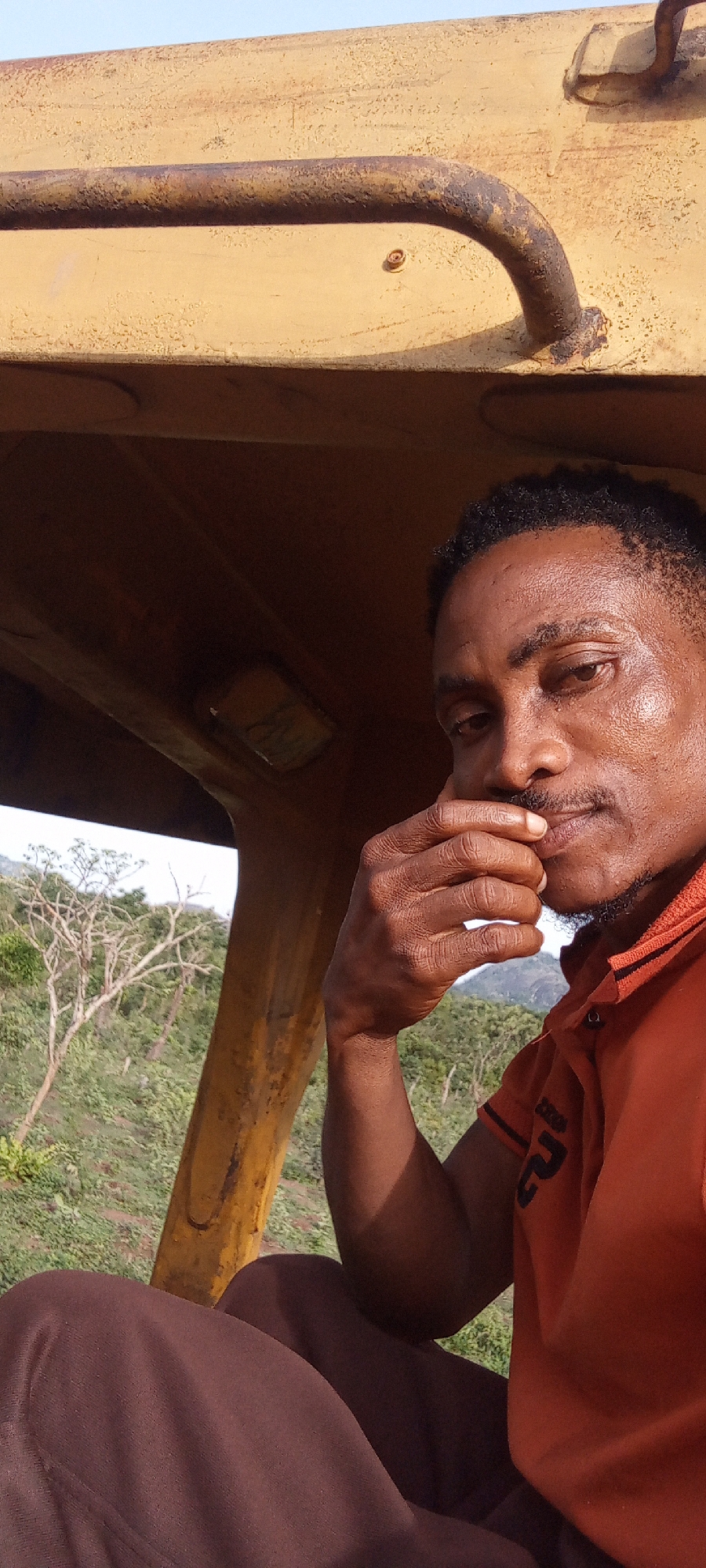 A man with dark skin and short, curly dark hair is depicted in a close-up, portrait-oriented shot. He is seated within the cab of a large, yellow machine, likely construction equipment, given the sturdy, weathered yellow metal frame and grab handle visible above and around him. He wears an orange collared polo shirt with black trim on the collar and sleeves, and a black stylized logo or text on the right sleeve. His lower body, covered in dark brown trousers, is partially visible at the bottom of the frame.
The man's right hand is raised, with his fingers resting gently near his chin and mustache, conveying a contemplative or thoughtful expression as he looks towards the camera. His face shows a slight sheen, suggesting warmth or recent activity.
Through the opening of the machine, a natural outdoor landscape is visible in the background. It features green foliage, some bare-branched trees, and distant hills or mountains under a bright, clear sky. This suggests a rural or undeveloped setting.
The time of day appears to be bright daylight with clear weather. Notable details include the rust and wear on the yellow metal parts of the machine, indicating its age and use. Inside the cab, above the man's left shoulder, a small, rectangular light fixture is visible with some barely discernible markings or text. The text on his shirt is stylized and difficult to read precisely.