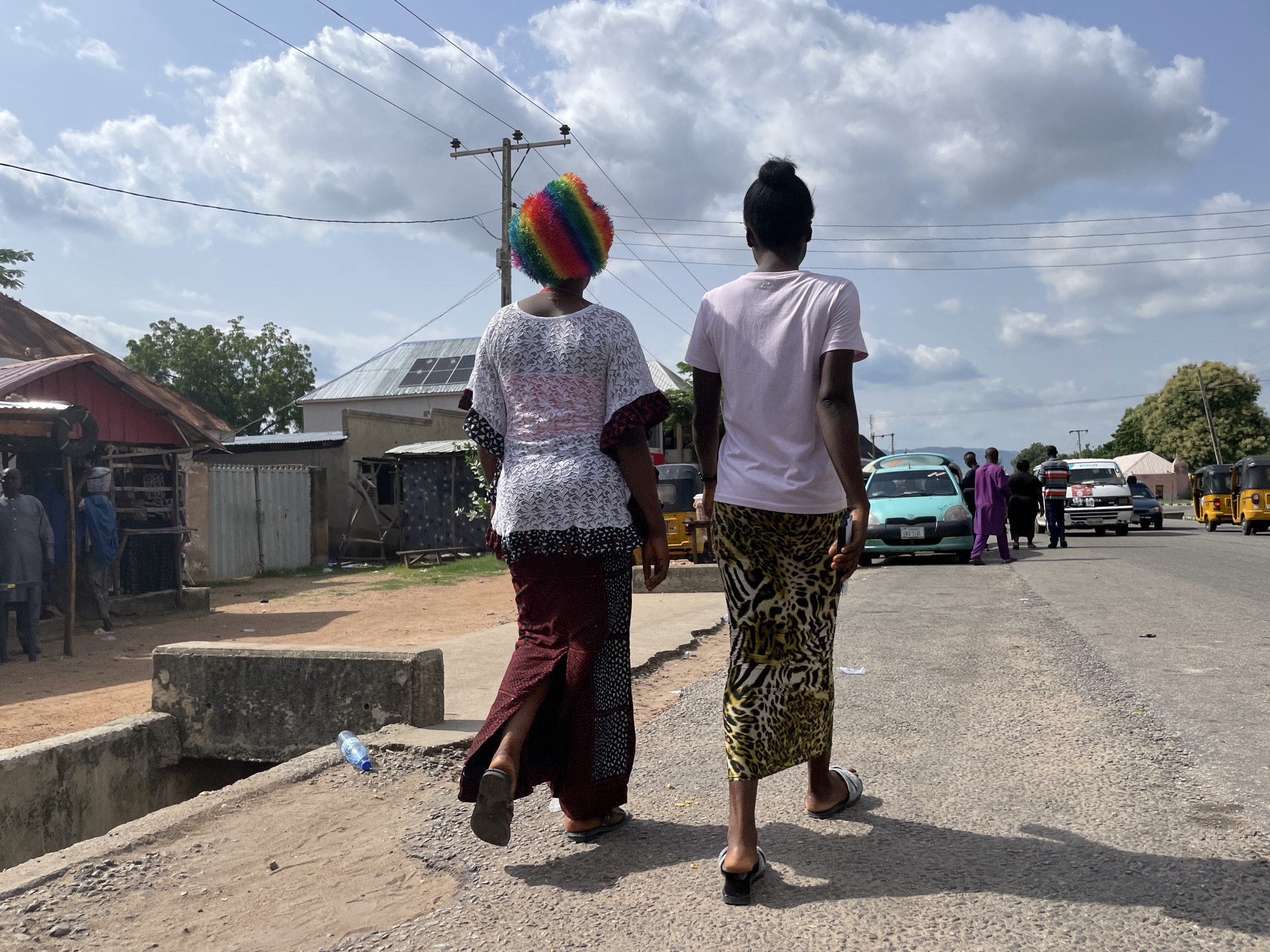 Two women are walking away from the camera on a paved road in Jalingo, Nigeria. The woman on the left wears a white lace top with a black and red patterned trim and a long, dark red and black patterned skirt. She also wears a colorful, rainbow-striped wig. The woman on the right wears a light pink t-shirt and a long, leopard-print skirt. She is wearing black sandals.
The road is asphalt and appears somewhat worn, with gravel visible on the edges. To the left of the road, there is a concrete drainage ditch with a bottle of water lying on its side near the edge. Behind the ditch, there are some structures that appear to be market stalls or small shops with corrugated metal roofs and walls. Trees and some buildings are visible in the background.
Several vehicles are present on the road, including a light blue car, a white van, and yellow motorized rickshaws (Keke Napep). Other people are also visible in the background, some near the vehicles and market stalls. The sky is cloudy with patches of blue. The scene suggests a typical day in a bustling area of the city.