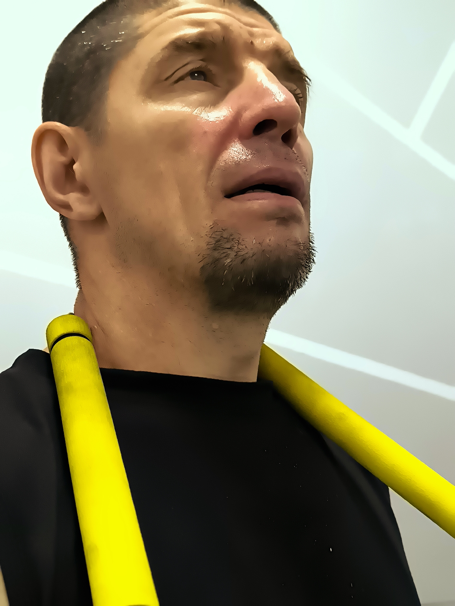 A man, likely of Caucasian descent, is depicted in a close-up, low-angle shot, looking upwards. He has short, dark hair, a shaved head on the sides and back, and a light stubble beard. His face is glistening with sweat, particularly on his forehead, temples, and upper lip, suggesting exertion or heat. His eyes are open, and his mouth is slightly ajar. He is wearing a black t-shirt, and a bright yellow cylindrical object rests across his shoulders, partially obscuring his collarbone and the upper part of his chest. The background is out of focus, with a light-colored wall featuring geometric white lines. There is no discernible connection to the city administration of Qarağandı, Kazakhstan.