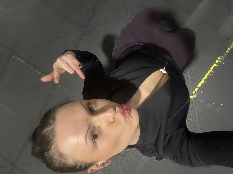A woman with fair skin, dark eyebrows, and dark hair pulled back is angled upward, puckering her lips. She wears a black, zippered athletic jacket, slightly open, revealing a white bra. Dark leggings are also visible. She holds up a hand with black nail polish, pointing down. The background is a dark gray, textured floor with a partially visible yellow line.