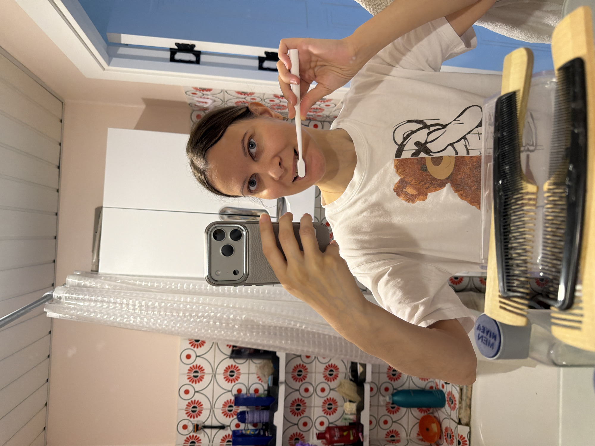 A young woman is in a bathroom, apparently in Izhevsk, Russia, taking a selfie while holding a white toothbrush near her mouth. She's holding a smartphone with a grey case. She is wearing a white T-shirt with a print of a bear. The background shows a white cabinet, tiled walls with a floral pattern, and a shower curtain. Two combs are visible on the right.