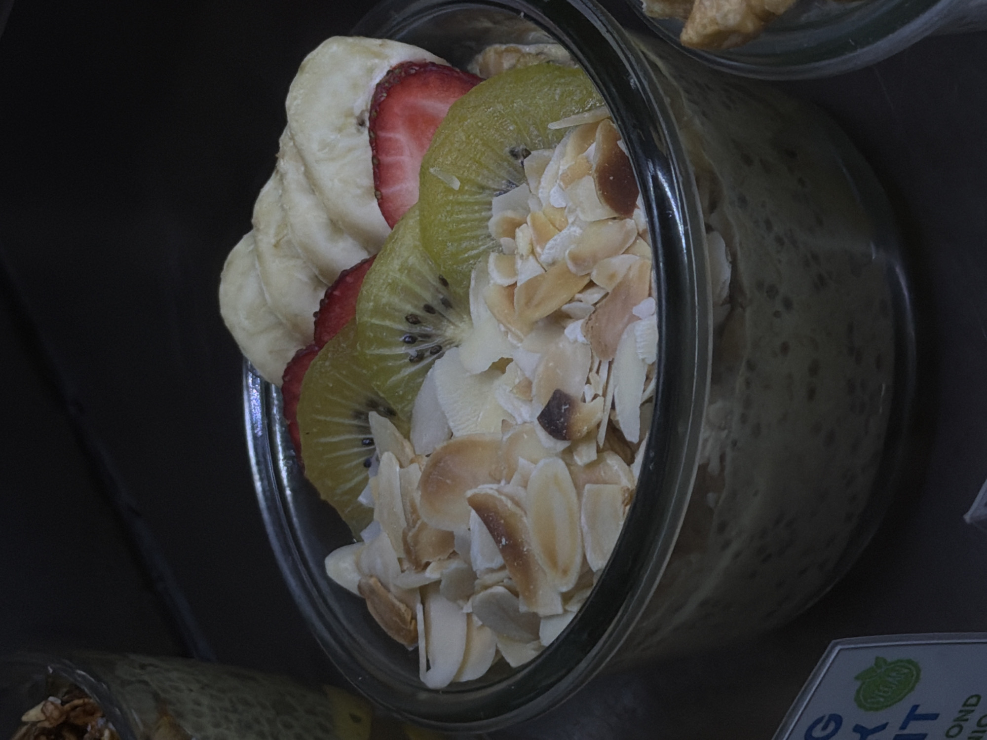 A close-up shot focuses on a clear glass bowl filled with overnight oats topped with sliced kiwi, strawberries, and slivered almonds. The oats appear to be a creamy, light yellow-green color with visible chia seeds or similar small particles. The fruit is arranged attractively on top, with the red of the strawberries contrasting with the green of the kiwi and the white of the almonds. In the background, a dark, out-of-focus area suggests a subdued lighting environment. To the lower left, a partial view of a sign with blue lettering and a green circular "Vegan" logo is visible. The scene suggests a cafe or food establishment. The image does not provide any specific information about the city of ตำบลเกาะพะงัน, Thailand, nor does it contain any people, actions, or interactions.