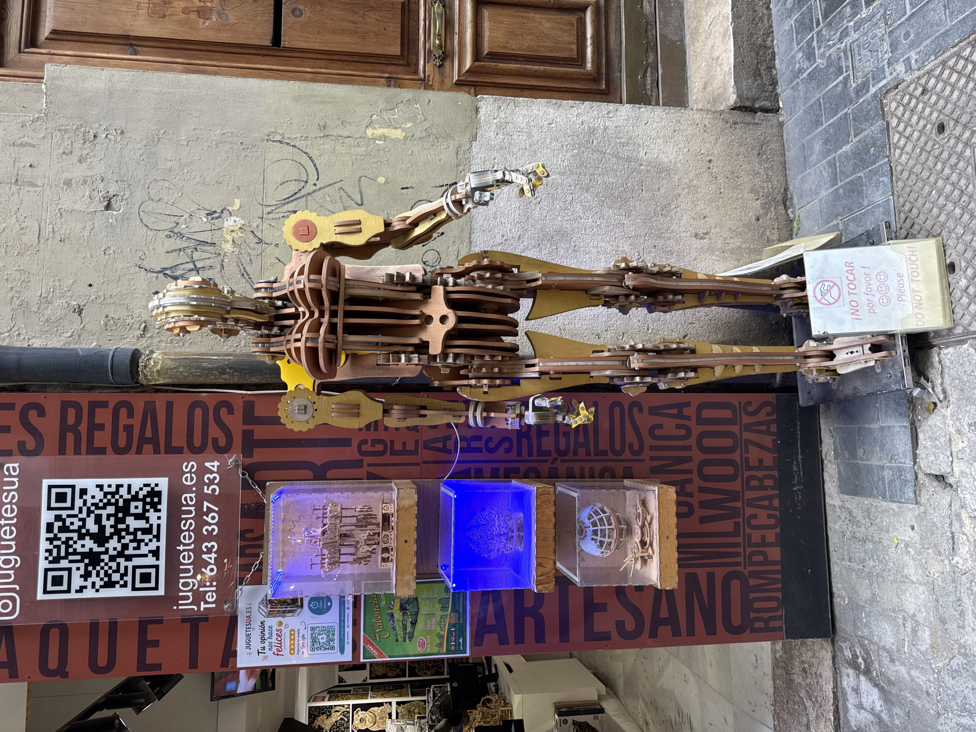 The image depicts the storefront of "JUGUETESUA" in Valencia, Spain. The shop specializes in wooden mechanical models and puzzles. A large, intricate wooden robot model stands outside the entrance, acting as a display or mascot. The robot is made of interlocking wooden pieces with visible gears and joints, and it is partially illuminated by ambient light.
A vertical banner to the left of the robot features a QR code, the shop's website "juguetesua.es", and a phone number "643 367 534". Below this, three transparent display cases are stacked vertically, each containing a detailed wooden mechanical model. The top case holds a carousel, the middle case displays a tree-like structure illuminated with blue light, and the bottom case contains a globe with gears.
Further to the left, part of the shop's interior is visible, showing shelves filled with various wooden puzzle kits and models. A sign with an image of a tram and the text "VALENCIA" is also present.
A white sign with red and black text is placed in front of the robot's base, reading "¡NO TOCAR por favor!" and "DO NOT TOUCH!". The signage and the name of the shop suggest it is a toy store. The background shows a textured wall with some graffiti and a wooden door to the right. The ground is paved with cobblestones and metal grates. The overall scene suggests an outdoor display area of a specialty shop in an urban environment.