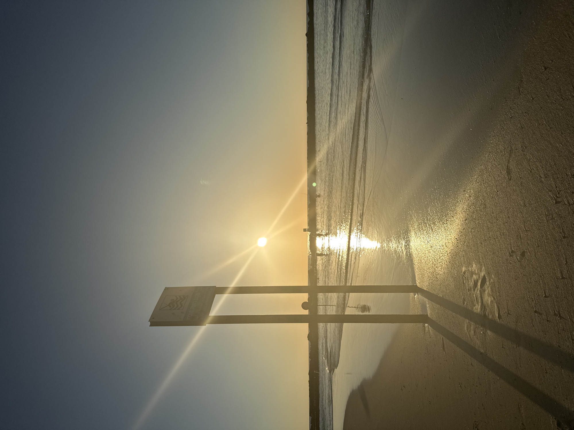 The image captures a serene sunset at a beach in Netanya, Israel. The sun, a bright orb, hangs low in the sky, casting a warm, golden glow across the scene. Rays of light fan out from the sun, creating a lens flare effect that adds to the dramatic atmosphere. In the foreground, a sandy beach stretches towards the water, with the wet sand near the shore reflecting the sunlight.
Dominating the left side of the image is a tall, thin signpost with a rectangular sign at the top. The sign, though partially obscured by the sun's glare, appears to have text and a graphic, possibly indicating rules or information about the beach. Two vertical poles support the sign, and their long shadows stretch across the sand, pointing towards the left. A small, circular object is attached to one of the poles, lower down.
The sea is calm, with gentle waves lapping at the shore. A breakwater, constructed from large rocks, forms a line across the horizon, separating the calmer inner waters from the open sea. A few figures can be seen in the water, wading or swimming, their forms silhouetted against the bright reflection of the sun on the water. The sky is a clear gradient of blue, transitioning from a pale yellow near the horizon to a deeper blue at the top. The overall impression is one of tranquility and natural beauty at the close of day.