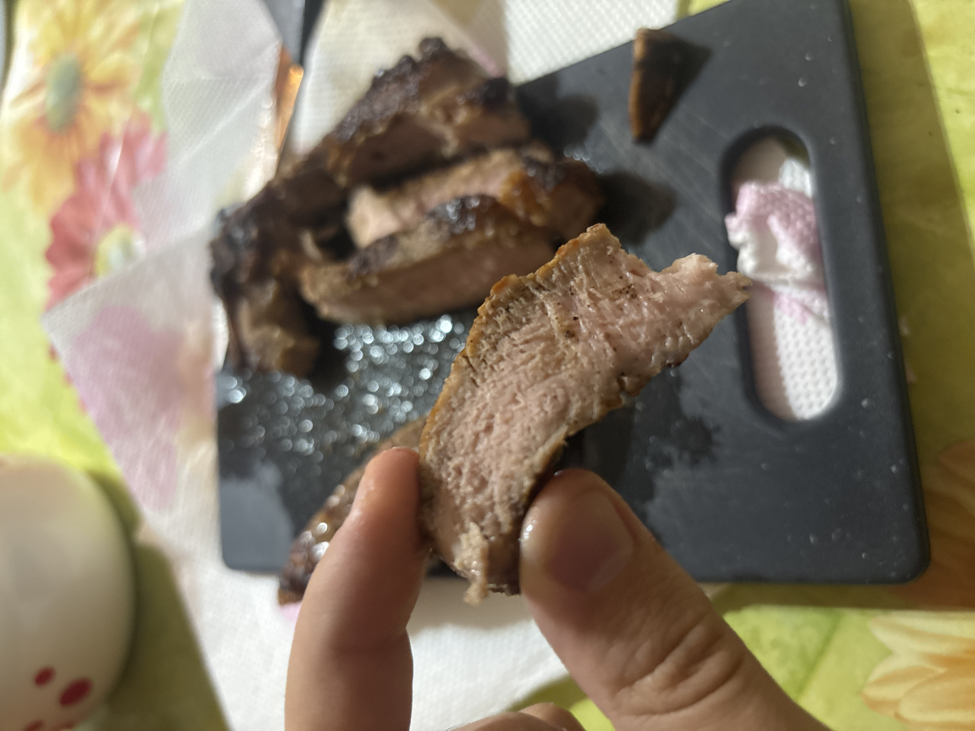 The image shows a hand holding a slice of grilled meat. More slices of the same meat are on a black cutting board in the background, which is resting on a white paper towel. The table under the paper towel has a floral design. The meat appears to be cooked medium-rare. There is not enough information to determine the city or location.
