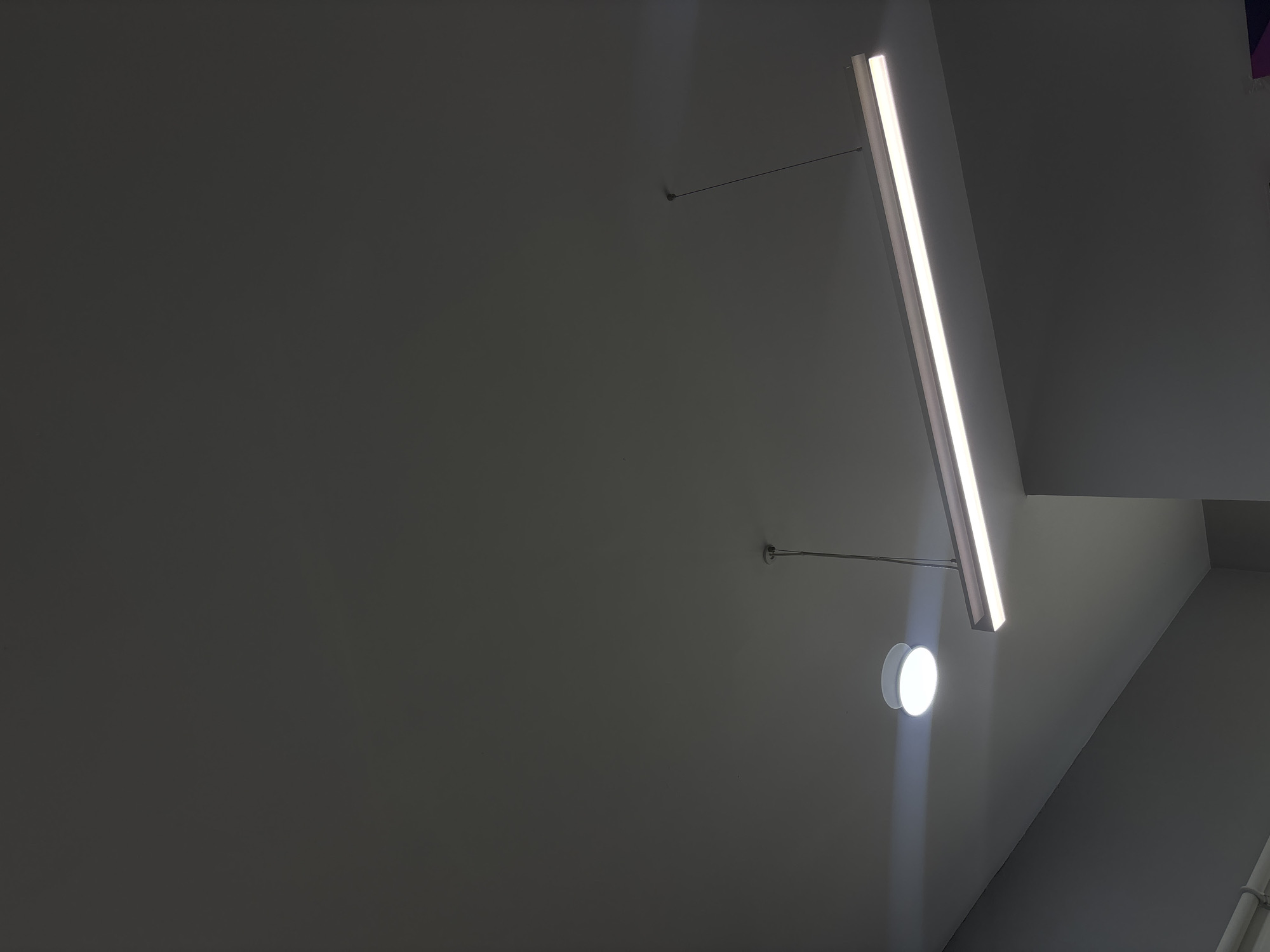 The image shows an indoor space with a gray wall and ceiling. Two light fixtures are visible: a long, linear LED light that is suspended from the ceiling by thin rods, and a round, wall-mounted light with a bright, focused beam. The wall is plain and without adornment. The space appears to be modern and utilitarian. The location in Buenavista, Mexico, is not discernible from the visual information in the image.