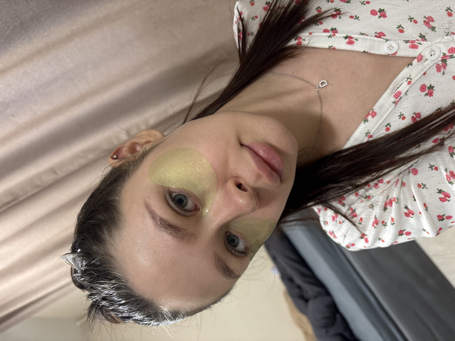 A woman with wet hair poses with gold eye masks. She has fair skin and blue eyes, wearing a floral white shirt with a silver necklace. The background features a beige curtain and grey fabric. Her face is slightly tilted, capturing a close-up view with her face centered. The photograph is well-lit, highlighting the skin and the masks.