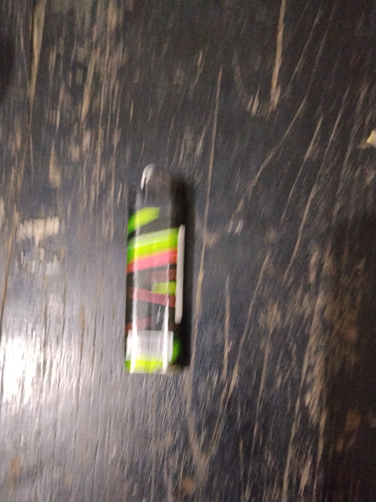The image shows a spray can with a black, green, and pink striped label lying on a dark, textured surface. The surface appears to be made of wood with prominent grain patterns and scratches. The spray can is positioned vertically. The lighting in the image is dim, creating a blurred and somewhat hazy effect. There is no information to suggest the location is in Bilis Bilis, Indonesia.
