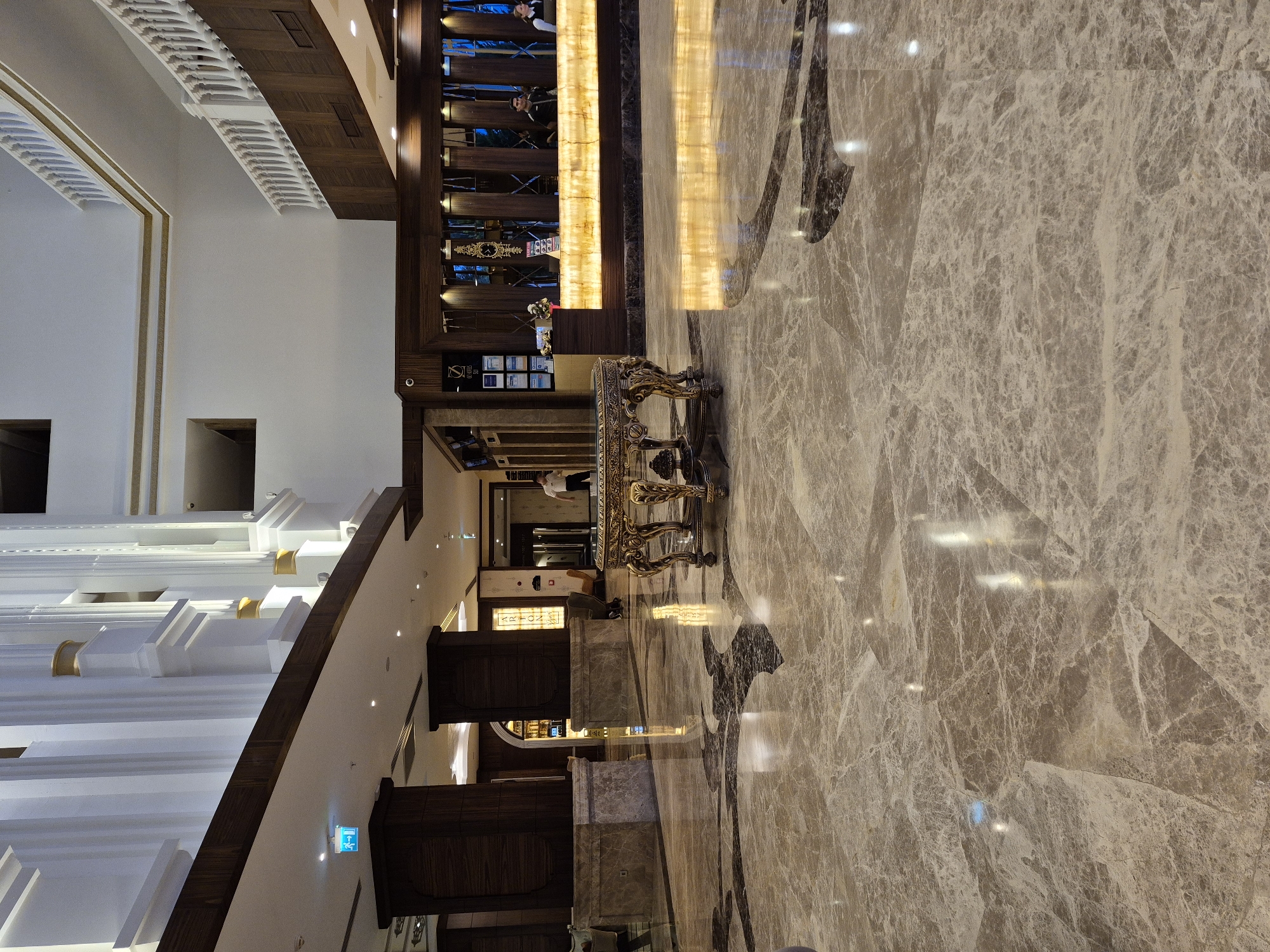 The image shows a section of a hotel lobby or grand hallway. The floor is covered in marble tiles with a mottled pattern, reflecting light from above. A gilded table with ornate legs stands in the center, and nearby is a reception or information desk. A wall panel is illuminated with a vertical strip of light behind the reception area. There are people standing by the reception desk. The architecture includes white columns with gold accents and wooden paneling on the upper walls. The overall impression is one of opulence and high-end design. The location is likely within the city of Alanya, Turkey, given the description provided.
