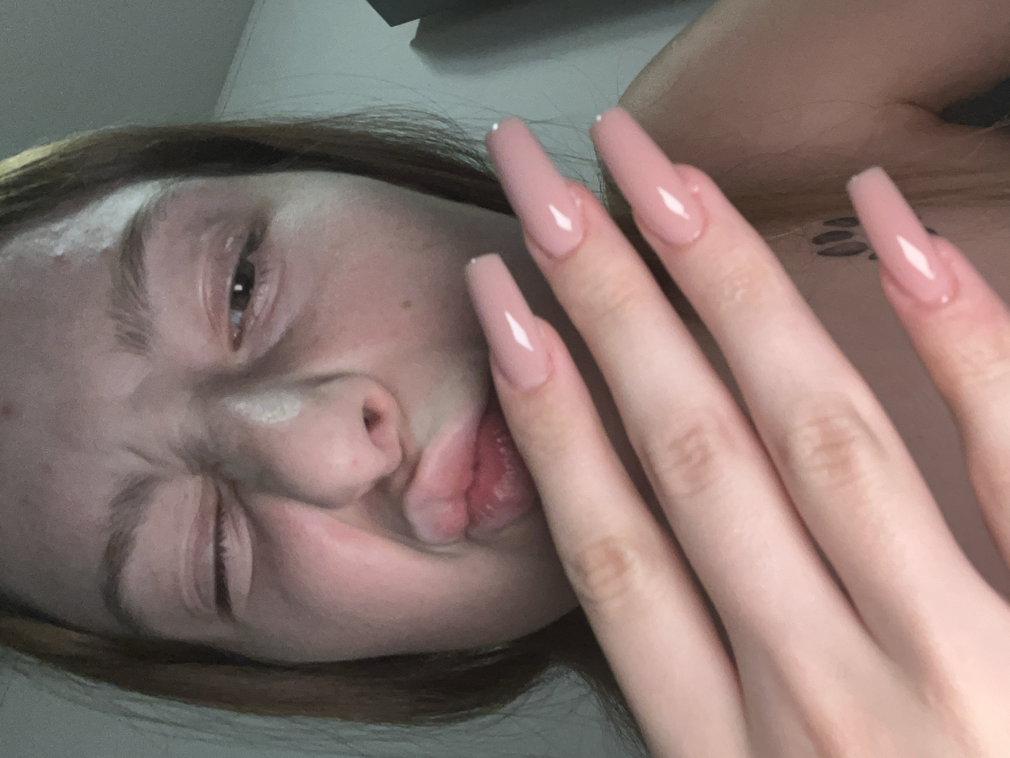 A close-up, off-kilter portrait shows a fair-skinned person with light brown hair. Their left eye is squinted shut, and their lips are pursed. Their hand, adorned with long, light pink, coffin-shaped nails, is positioned near their face. The background is muted and out of focus. A small tattoo is visible on their chest. There is no contextual information to suggest a location in Varna, Bulgaria.