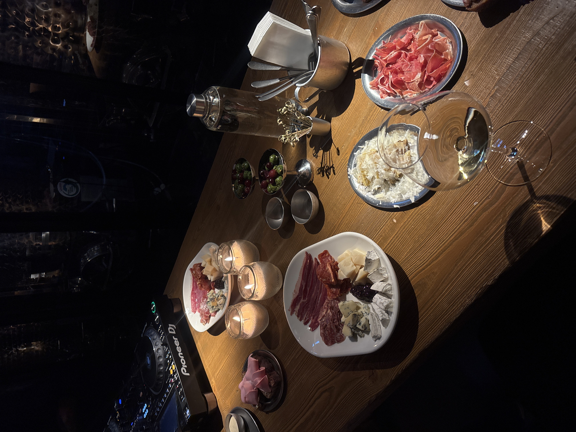 The image shows a high-angle view of a wooden table in a dimly lit setting. The location is unknown, although the prompt suggests Moscow, Russia. The table is set with various plates and serving dishes, including a charcuterie board with meats and cheeses, a plate of ham, a plate of shaved parmesan, a dish of olives, and a cocktail shaker with cocktail forks and napkins. There are also several small candle holders with lit candles, and two wine glasses, one filled. In the lower left corner, there is a Pioneer DJ controller, suggesting music entertainment is present. There are no people visible. The scene suggests a social gathering with food and drinks.