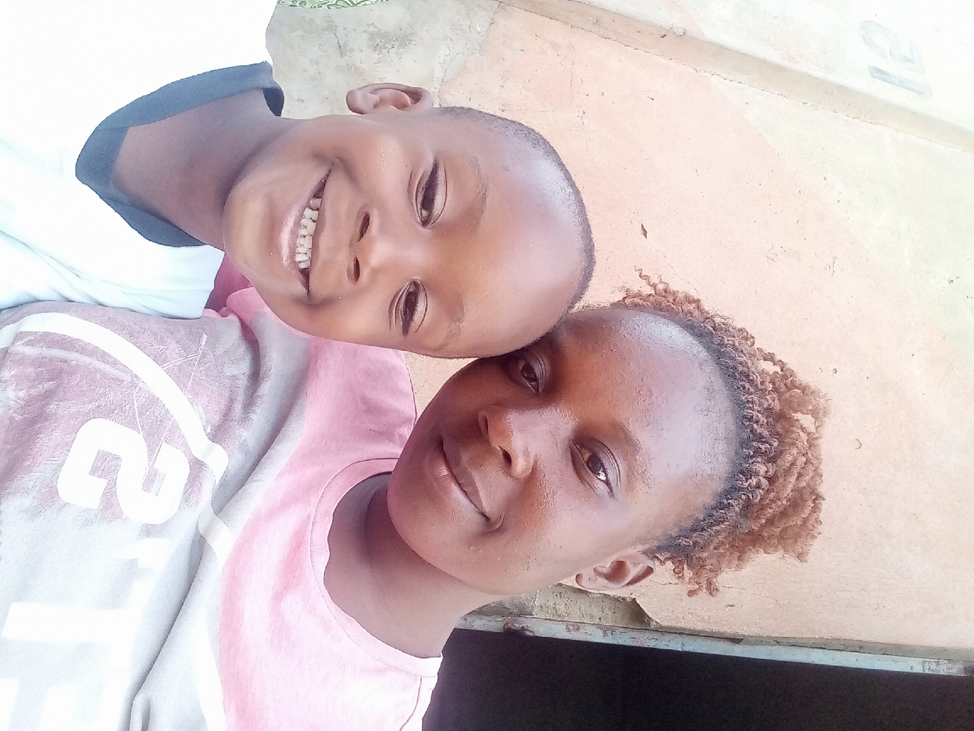 A close-up, slightly rotated, selfie-style image shows a woman and a child. The child, who appears to be a young boy, is in the top left corner of the frame, smiling broadly with his teeth visible. He has short, dark hair and wears a white shirt with a dark collar. His head is positioned on or very close to the woman's head. The woman has dark skin, short curly hair, and a gentle smile. She wears a pink t-shirt with a white design on it. The background is out of focus, but a beige or light brown surface is partially visible, possibly a wall. The location is Sabon Tasha, Nigeria.