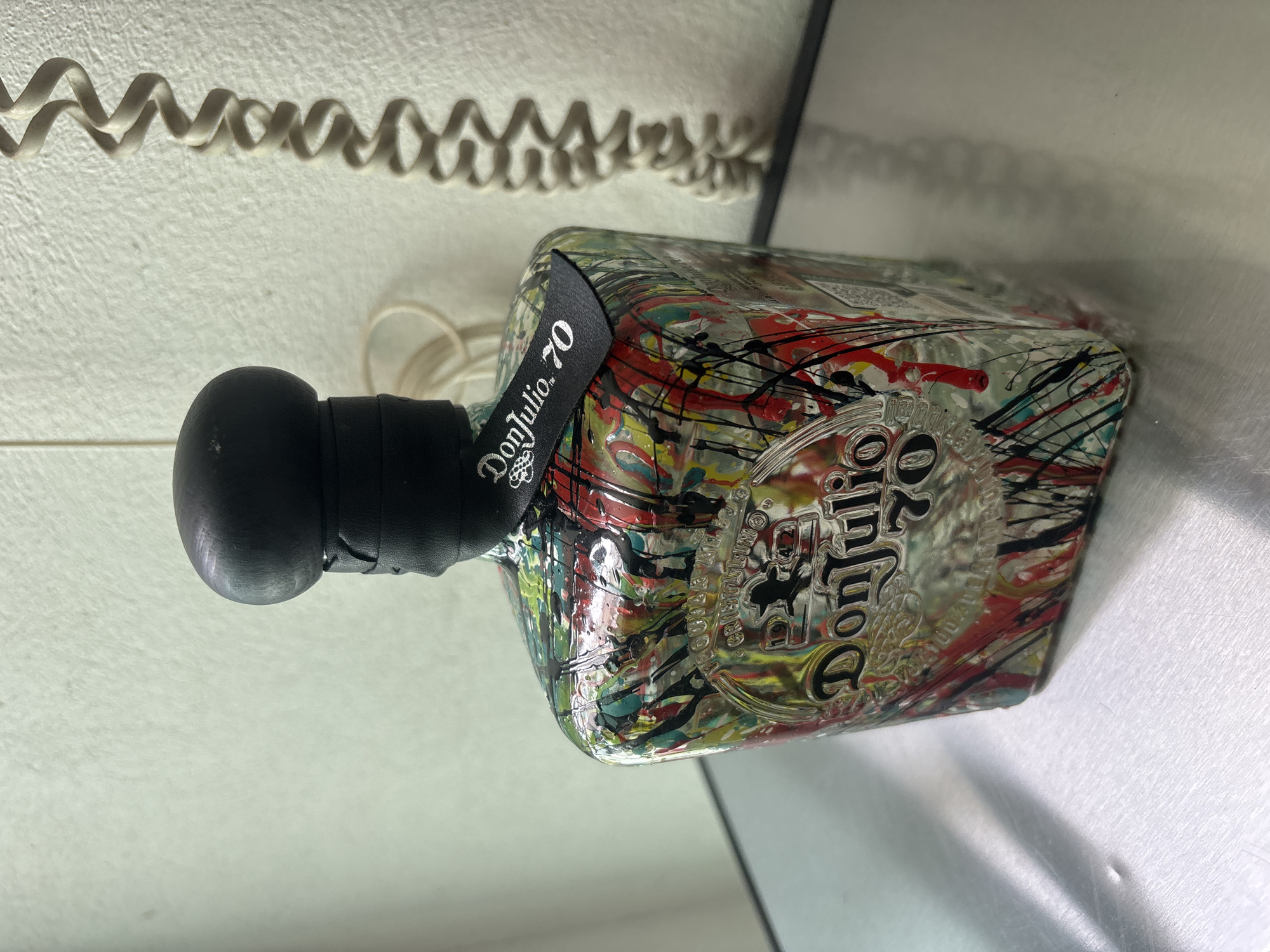 The image shows a bottle of Don Julio 70 tequila lying horizontally on a surface. The bottle has a black cap and neck label. The glass is decorated with colorful paint splatters. A coiled cord is visible in the background. The image provides no information to confirm the location is in Jesús del Monte, Mexico.