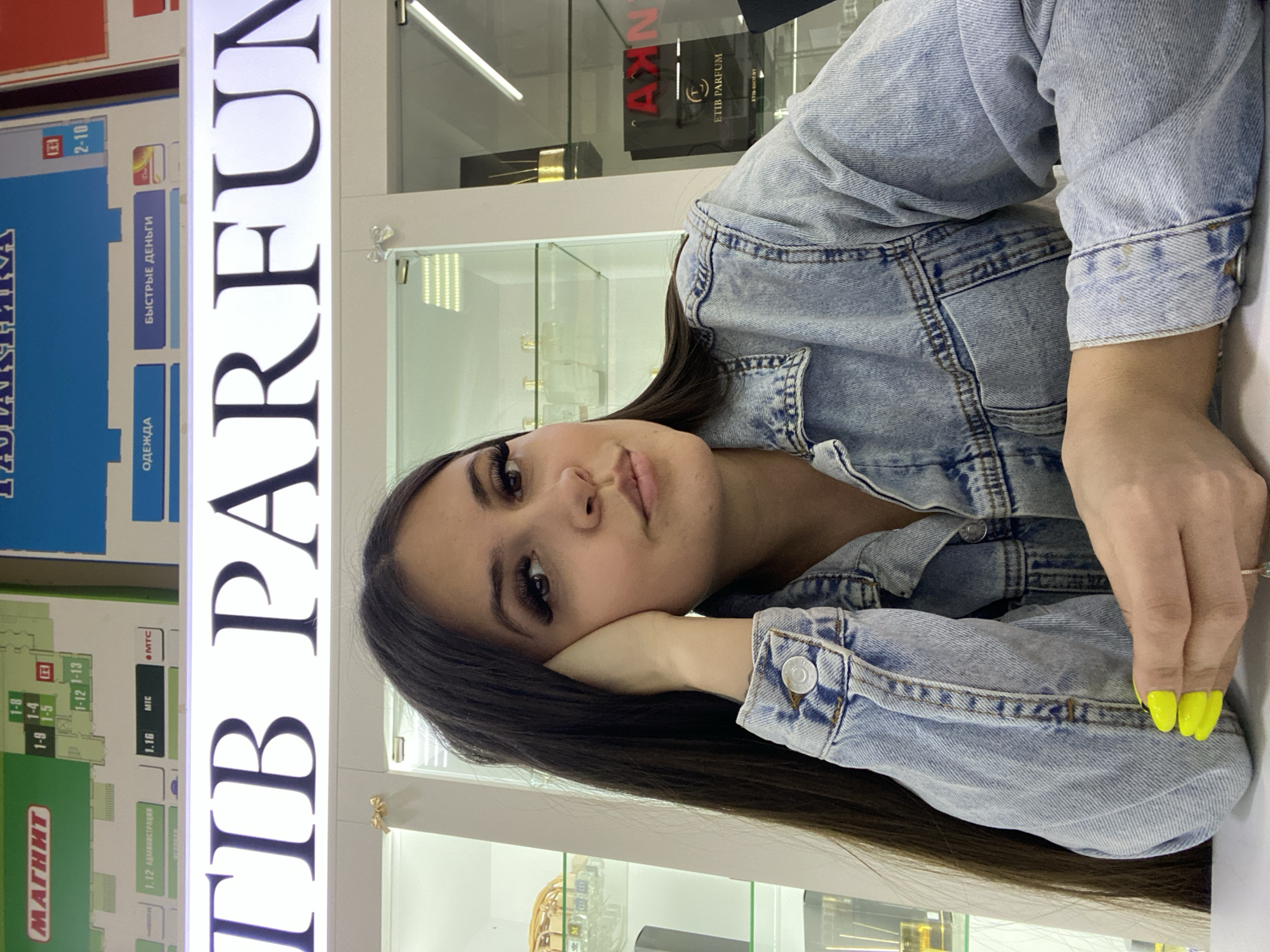 A young woman with long, dark hair is photographed indoors, resting her head on her hand. She is wearing a light blue denim jacket. Her nails are painted bright yellow. Behind her is a display with signs written in Cyrillic, including the words "TIB PARFUM". Signs also indicate "MAGNIT," a Russian supermarket chain. The image includes text indicating "быстрые деньги" (fast money) and various floor numbers. One sign is labeled "ETIB PARFUM." Based on the signs, the location appears to be a shopping center or mall in Chaikovsky, Russia.