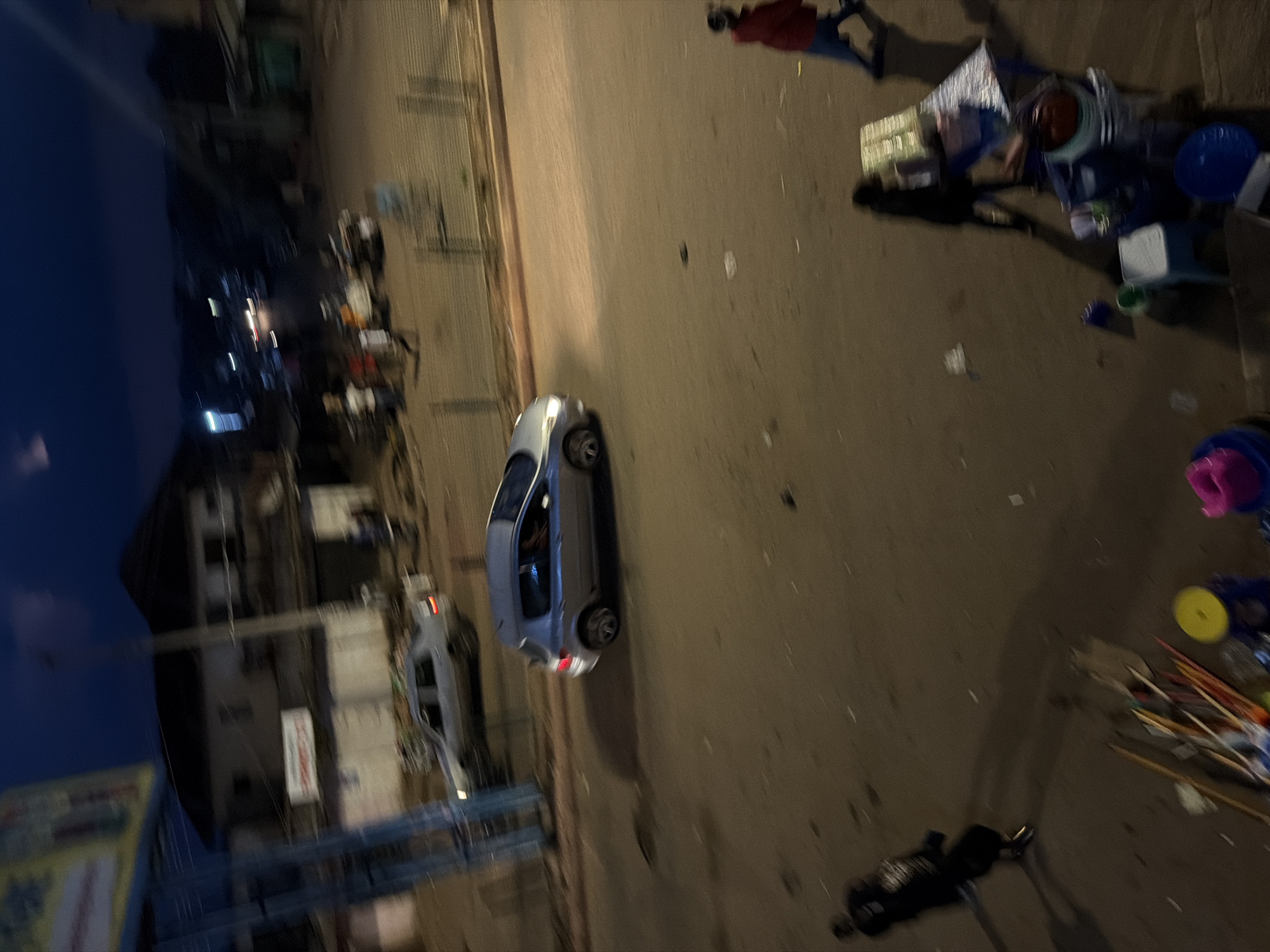 The image depicts a street scene at dusk or night in Bukuru, Nigeria. A silver hatchback car is driving on the right side of the road, with its headlights on. Behind it, a lighter colored sedan is also visible. Several people are on the street, some walking and others standing near what appear to be market stalls or vendors. There are various objects scattered on the ground, possibly debris or goods for sale. Buildings are visible in the background, illuminated by streetlights. The overall lighting suggests low light conditions, characteristic of evening or nighttime.