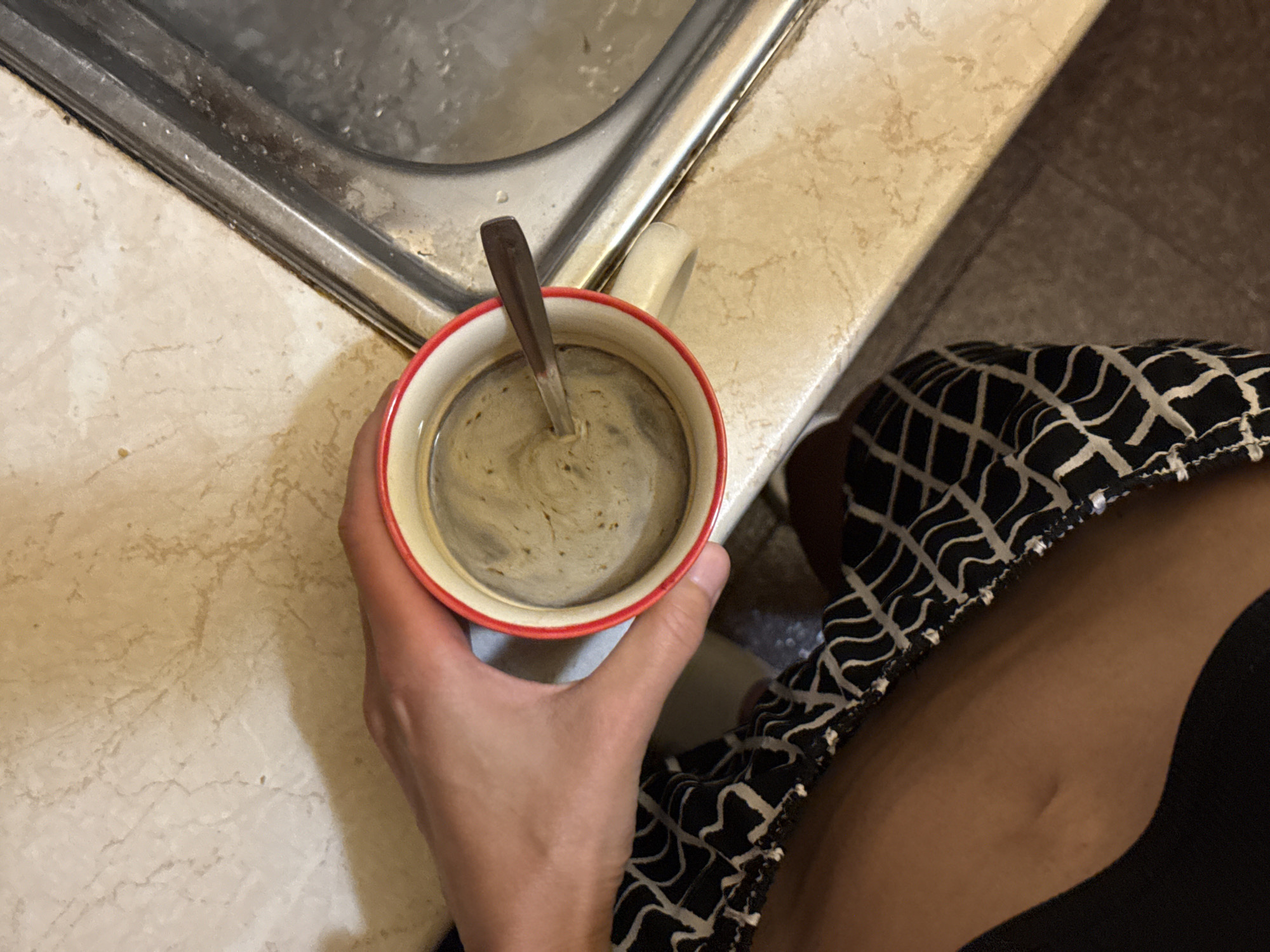 A person's hand holds a white mug with a red rim, containing coffee. A spoon rests in the mug. The mug is positioned on a countertop with a marble-like pattern. To the right of the mug is a stainless steel sink. The lower portion of the image shows a person's legs clad in black and white patterned pants. The scene appears to be an indoor setting, likely a kitchen or bathroom. No specific location in Pleven, Bulgaria is identifiable from the image.