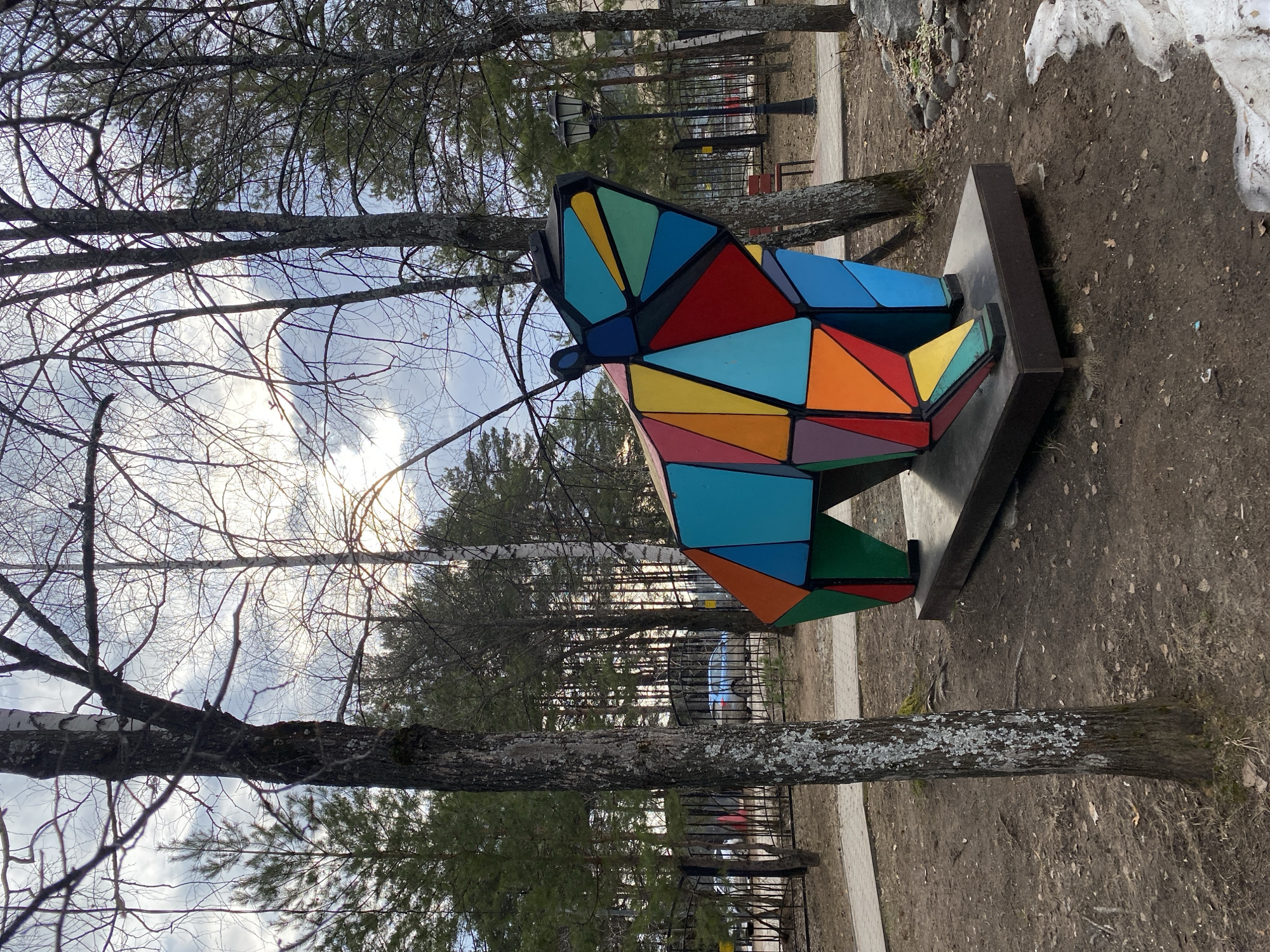 The image, taken in Chaikovsky, Russia, features a vibrant, geometric sculpture of an animal, likely a fox or a dog, situated outdoors. The sculpture is composed of numerous triangular and polygonal panels in a mosaic of colors including blue, red, orange, yellow, green, teal, and purple, all mounted on a dark, rectangular base.
The scene depicts a park-like setting during daytime, likely in late winter or early spring. This is indicated by the bare branches of the deciduous trees, the mostly dirt ground with sparse dry grass, and patches of melting snow near the edge of the frame. The sky above is overcast, with bright patches suggesting the sun is diffused behind the clouds.
Numerous trees with dark, textured bark and intricate, leafless branches dominate the midground and foreground, creating a natural frame. Some evergreen trees are also visible in the background. A paved path runs through the scene, and a black metal fence can be seen further back, behind which a street lamp and a blue car are partially visible. There is no discernible activity taking place, and no text is visible in the image.