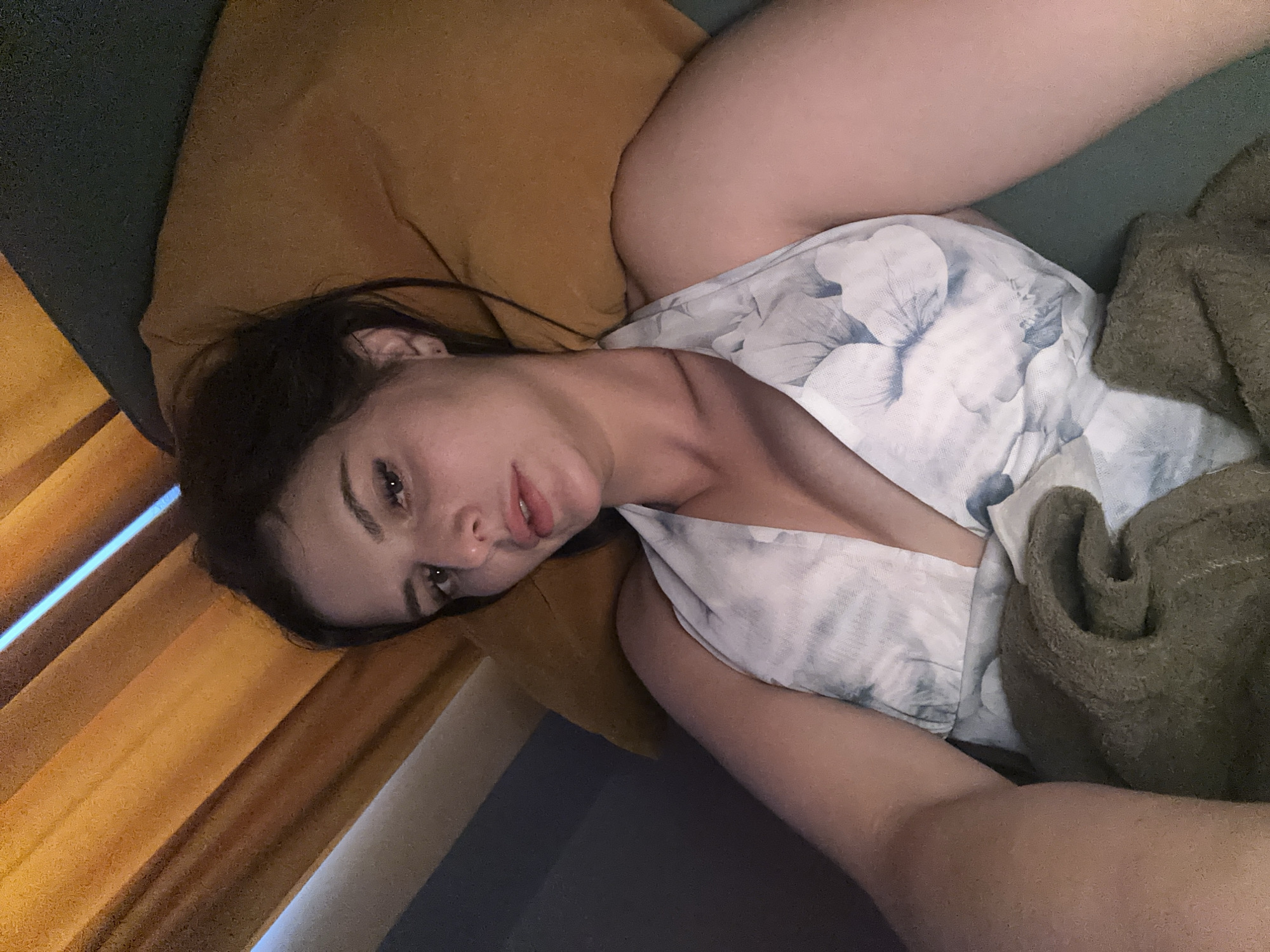 A woman with dark hair is lying down, her head resting on a tan pillow. She is wearing a white halter top with a floral pattern. Her eyes are open and looking towards the camera, and her mouth is slightly parted. A brown blanket is draped over her. The background appears to be an indoor setting, with wooden slats and a blue light source visible above the woman. The scene is lit with warm, artificial light. The location is unknown.
