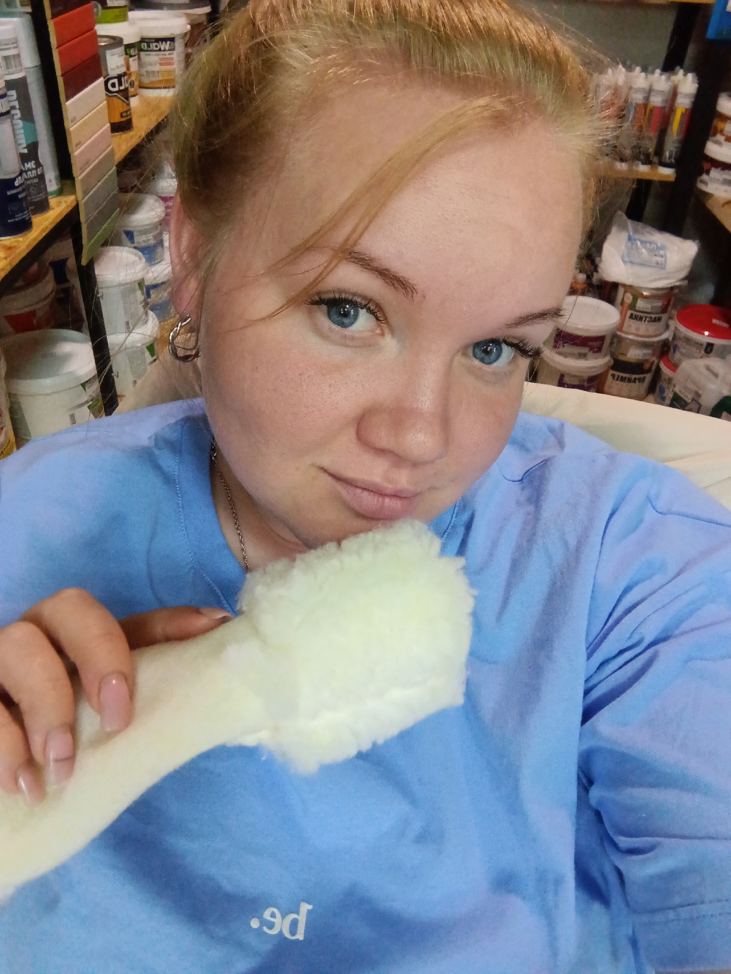 A fair-skinned, blond-haired woman with blue eyes is holding a cream-colored, fluffy object. She is wearing a light blue t-shirt. The background shows shelves filled with containers and paint supplies, suggesting a store environment. A location, Фоки, Russia, is mentioned, but there is no visual indication within the image to confirm or deny this location.