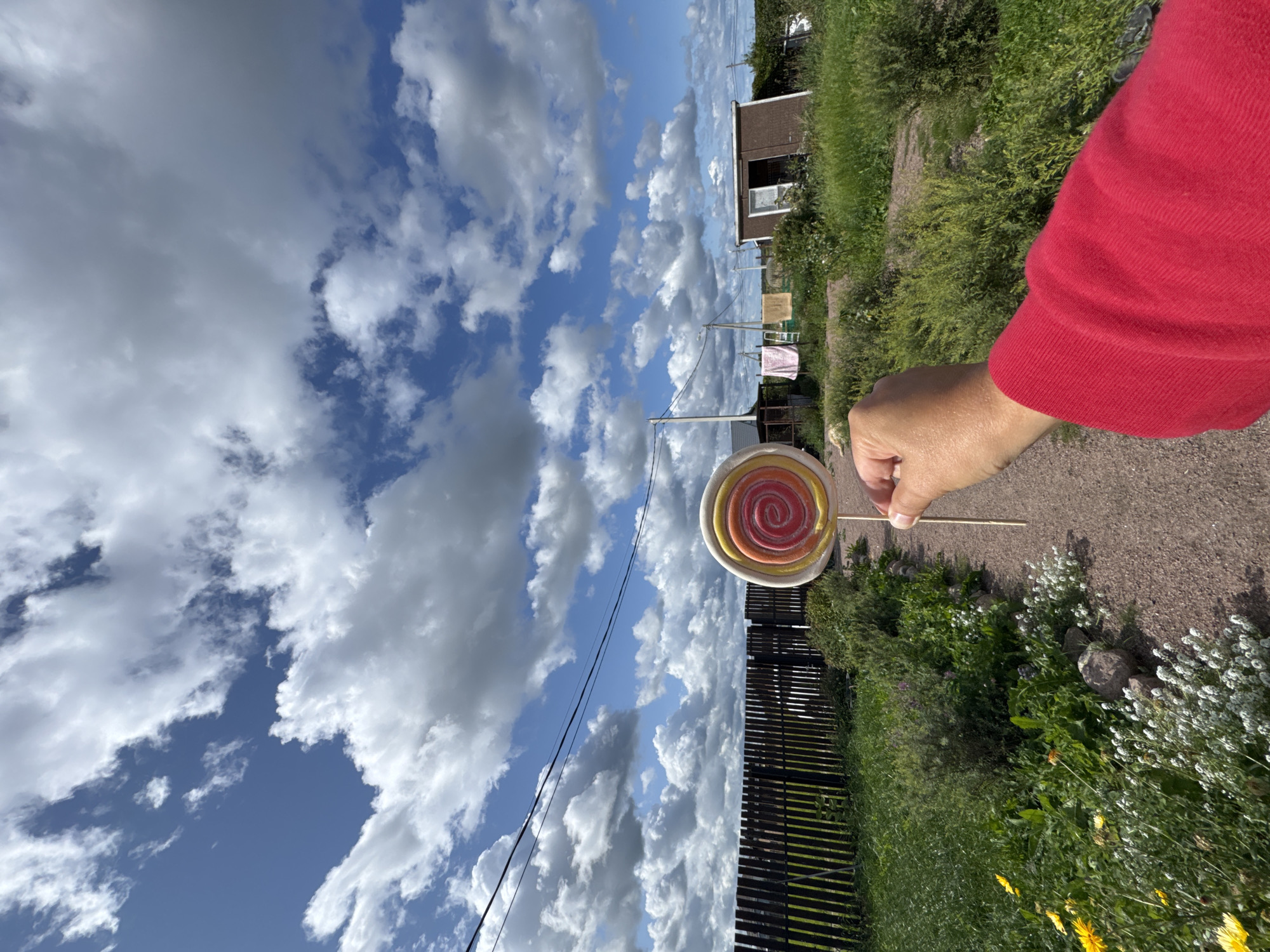A person wearing a red long-sleeved shirt is holding a large, multi-colored spiral lollipop in their right hand, in the foreground of the image. The lollipop is pink, yellow, and white. The background features a rural landscape in Kempelevo, Russia. A dark wooden fence runs horizontally across the middle ground, with green grass and various plants in front of it. Beyond the fence, there are several small buildings, including a brown shed with an open door, and laundry hanging on a clothesline. Power lines are visible stretching across the bright blue sky, which is filled with dramatic white and gray clouds. A gravel path leads away from the viewer towards the fence.