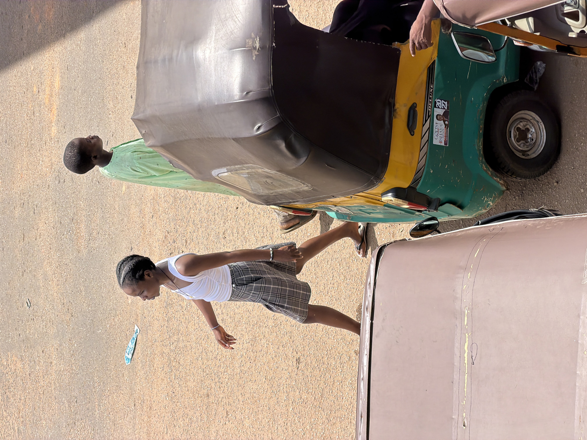 In Bukuru, Nigeria, on a sunlit, dusty road, two young individuals are observed walking. A young girl, wearing a white tank top and a grey plaid skirt, walks from left to right, her gaze directed forward. Behind her, a person in a light green garment walks in the same direction. In the foreground, the rear of a brown vehicle is visible, and to the right, a green and yellow auto-rickshaw, with a black canvas top, is parked. The auto-rickshaw has stickers on its side, one featuring a portrait and the text "GOD'S ISHAYA 2022". The road surface is predominantly sandy and light brown.