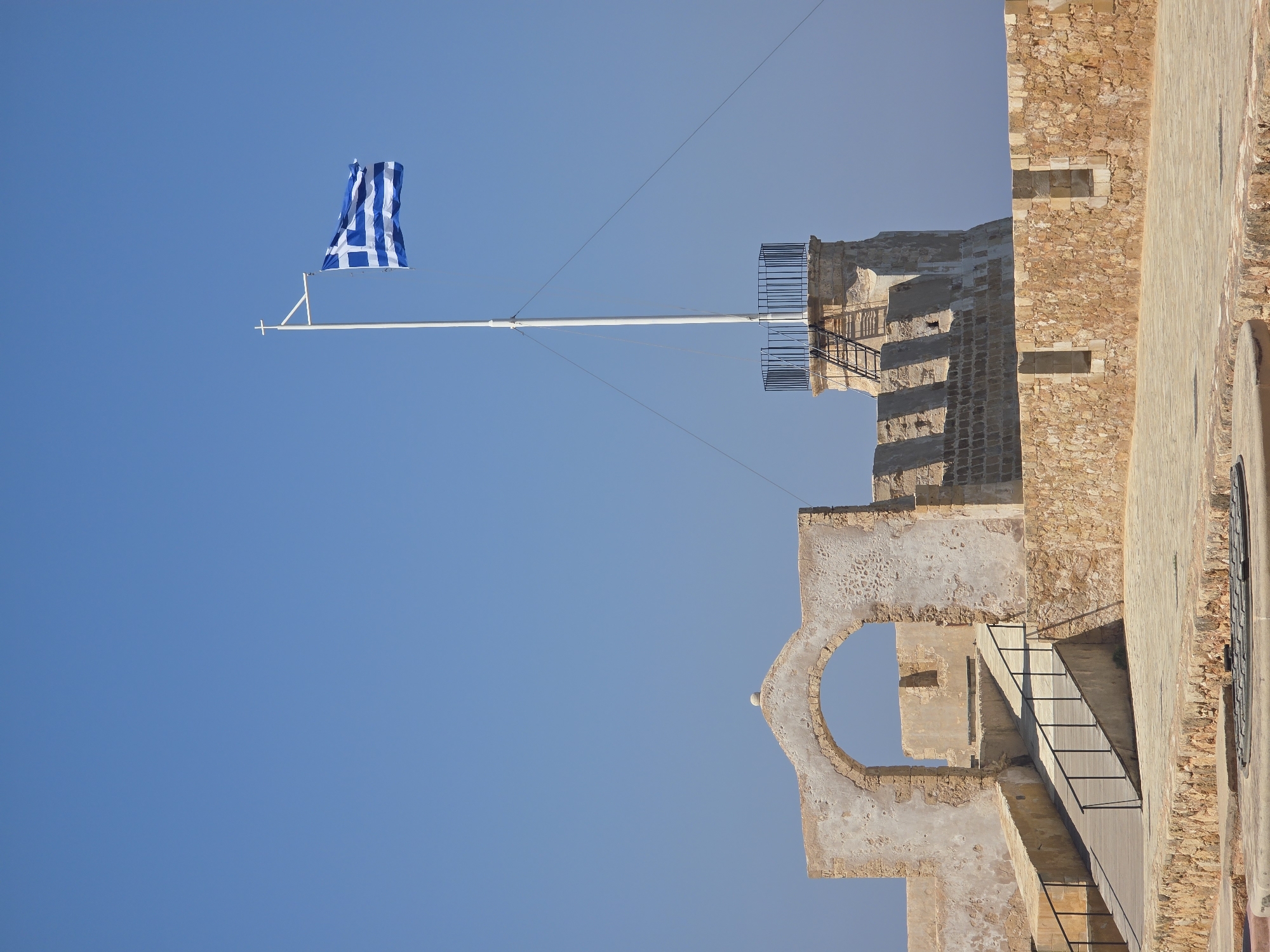 The image depicts the Fortezza of Rethymno, a Venetian-era fortress situated in the city of Rethymno, Crete, Greece. The scene is dominated by the clear blue sky and the weathered stone walls of the fortress. A Greek flag flutters from a tall flagpole, positioned atop a cylindrical tower that is part of the fortress. In the foreground, a stone pathway leads towards an arched gateway, with a wooden ramp and metal railings on the left. The fortress walls are constructed from rough-hewn stone blocks, with visible signs of age and erosion. The overall impression is one of historical significance and a sense of place, typical of ancient fortifications in the Mediterranean region. There are no people visible in the image.