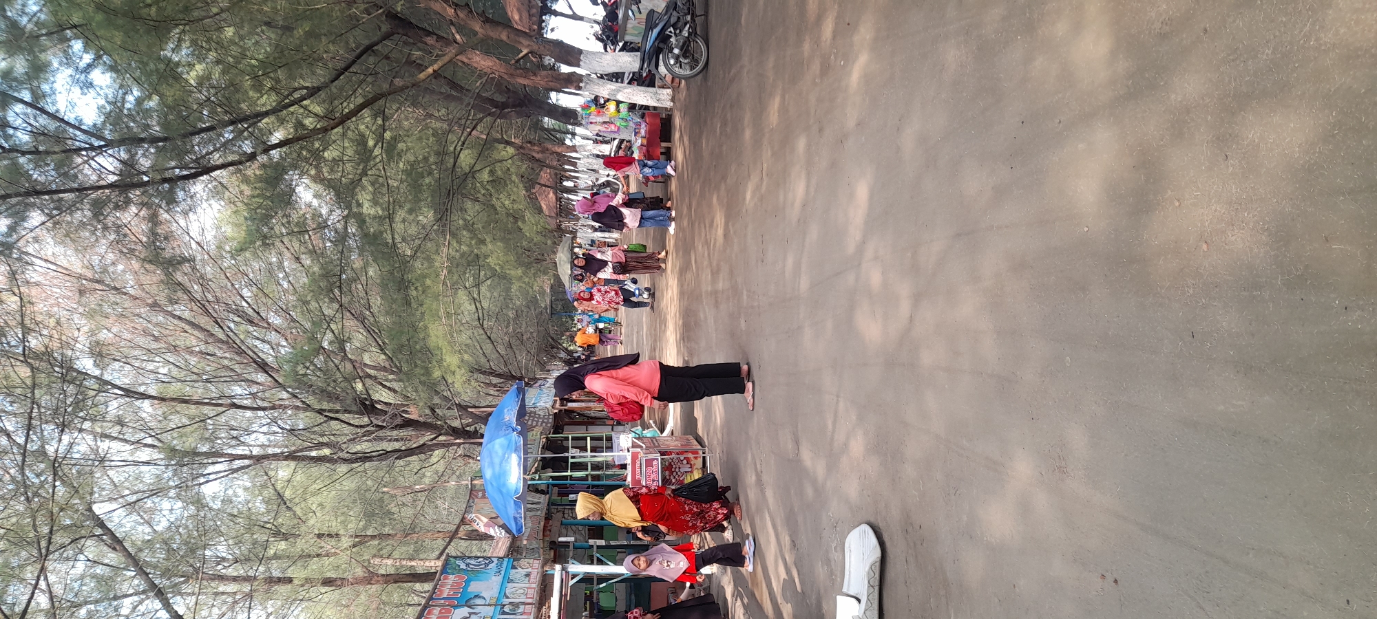 The image depicts a bustling pathway lined with trees and market stalls, likely within the Nyamplung Sari area of Indonesia. The ground is a dusty, sandy surface, with a few scattered leaves and debris. On the left side of the pathway, there are several market stalls, one with a blue umbrella. Various items are displayed for sale, including food and trinkets. Several individuals, predominantly women wearing hijabs and long dresses or pants, are present. Some are browsing the stalls, while others are walking along the pathway. In the background, more people and stalls are visible, suggesting a lively market environment. Trees with thin branches and sparse foliage form a canopy overhead, dappling the pathway with sunlight. A motorcycle is parked on the right side of the pathway, near some stalls that have white-painted bases. A white sandal is visible on the lower left of the image. The overall scene suggests a vibrant marketplace located in a coastal or beachside area.