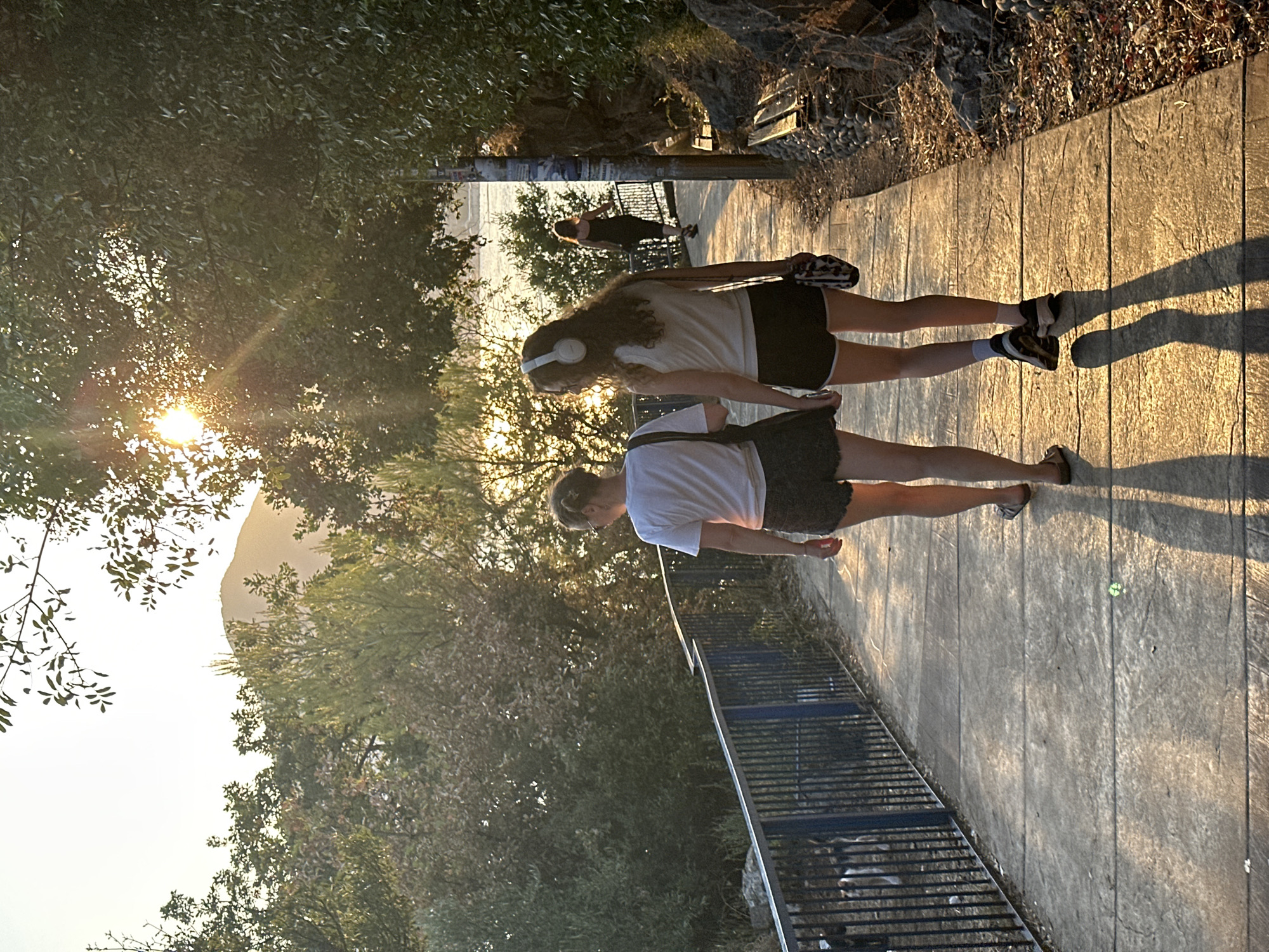 Two young women walk away from the camera on a paved pathway that runs alongside a blue railing. The woman on the left wears a white t-shirt and dark shorts, her hair pulled back in a bun. The woman on the right wears a white tank top and black shorts and has white headphones on. Behind them, further down the path, a third woman in a dark dress walks away from the camera. The pathway is lined with trees and rocks, and the sun is setting, casting long shadows. The scene appears to be a scenic overlook or park area. The location is believed to be in Budva, Montenegro, likely near the coast given the presence of water and lush vegetation.