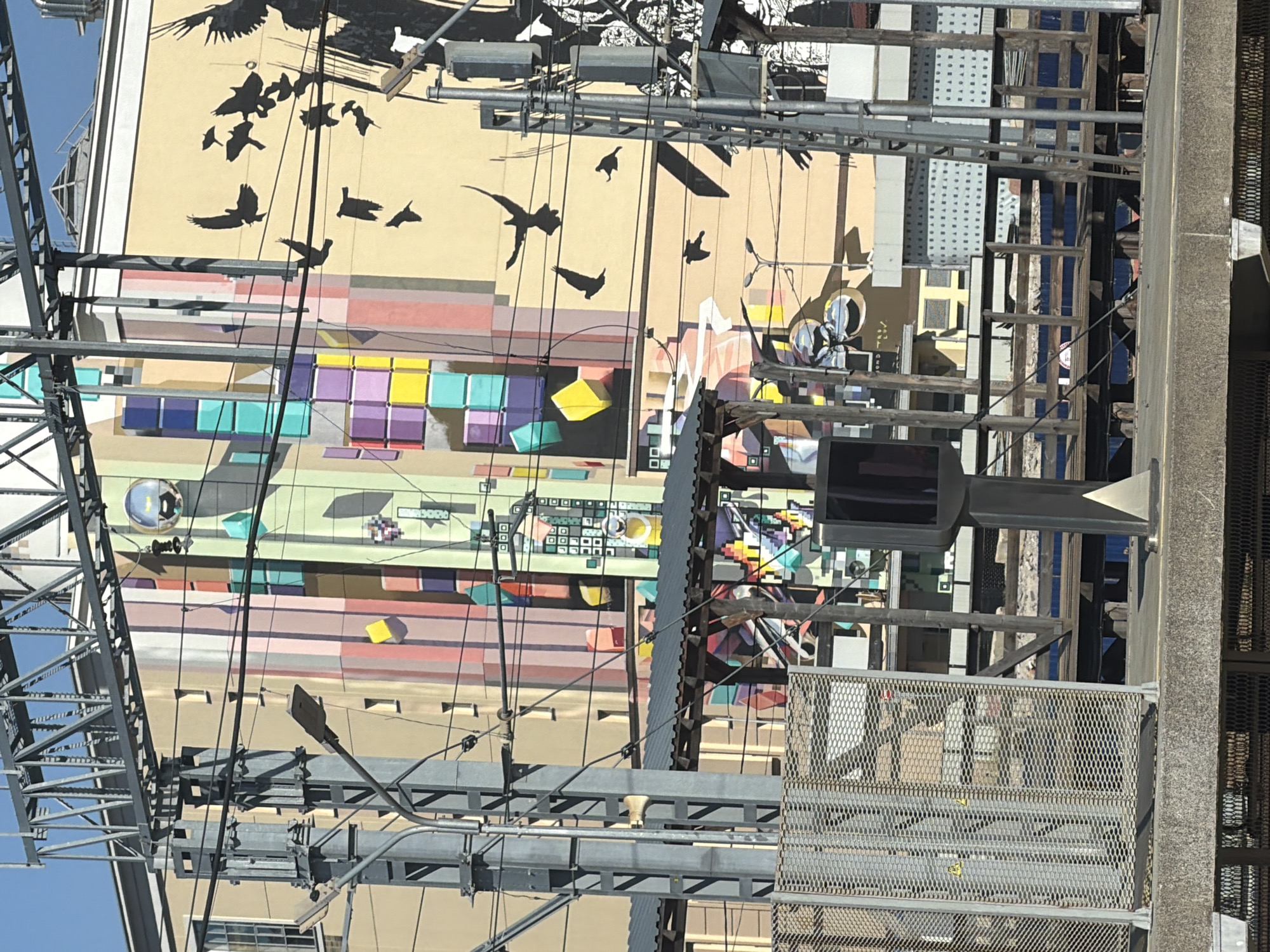 A mural depicting geometric shapes and silhouetted birds adorns the side of a building, partially obscured by overhead train lines and infrastructure. The building facade itself is painted in stripes of various colors, including yellow, pink, and green, overlaid with a mural that features black silhouettes of birds in flight and abstract colorful blocks. Below the mural, a covered walkway with a corrugated metal roof is visible, supported by wooden beams and metal poles. In the foreground, a gray metal fence with a honeycomb pattern encloses an area, and a traffic signal stands to the right of the fence. The scene appears to be a transportation hub, likely a train station, given the presence of tracks, overhead wires, and a platform. The architectural elements and style do not suggest Moscow, Russia.