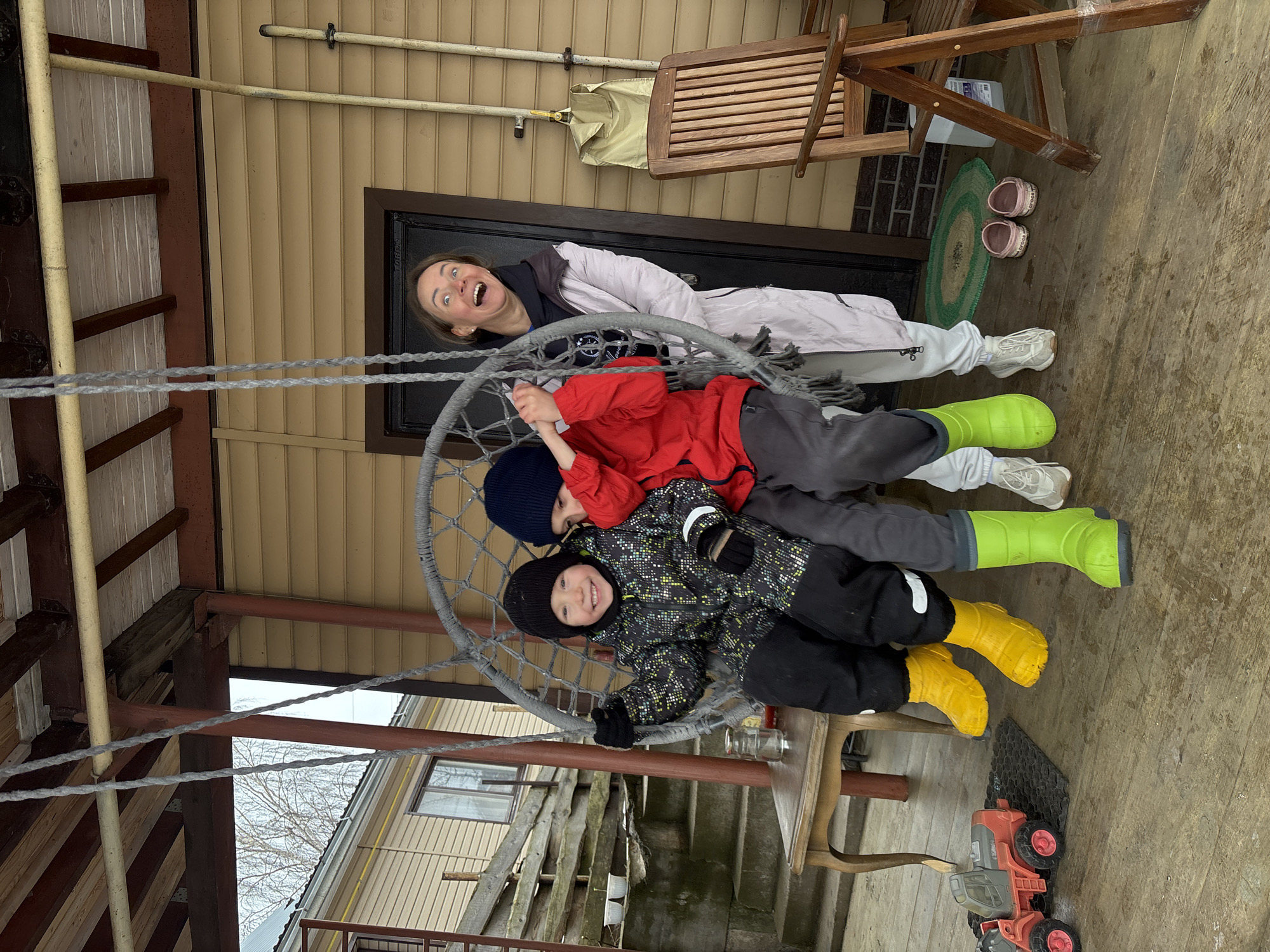 The image captures a candid outdoor scene. A woman lies horizontally near a door in a beige-colored building. The woman is making a silly face, her mouth wide open. Below her, two young children are seated together in a hanging circular chair, or swing. The child on top is wearing a red jacket, grey pants, and bright green rubber boots. The child below is wearing a patterned dark jacket, black pants, and yellow rubber boots. Both children wear dark knit hats. Below the swing, on the wooden flooring, is a toy tractor. The setting appears to be a covered porch or patio of a building in a residential area. Based on the user's provided location, the scene is occurring in Chaikovsky, Russia.