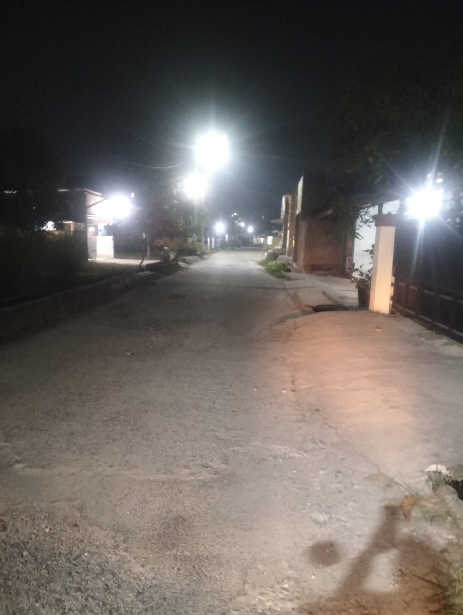 The image depicts a dimly lit street scene at night in Air Jamban, Indonesia. The street is paved with asphalt and appears to be a residential or semi-commercial area. Several streetlights illuminate the scene, casting long shadows and creating areas of bright light and deep darkness.
On the left side of the street, there is a building with a light-colored awning, possibly a small shop or eatery, with visible lights inside. Adjacent to this building are trees and vegetation. Further down the street, more lights and buildings are visible.
On the right side of the street, there is a tall, light-colored pillar and a dark-colored gate or fence. A potted plant is visible near the pillar. The ground on the right side of the street shows a lighter, smoother surface compared to the rougher asphalt of the road.
The overall impression is that of a quiet, possibly suburban street after dark, illuminated by artificial lighting. There are no people or vehicles visible in the frame, suggesting a low level of activity. The image is taken from a low-angle perspective, looking down the length of the street.