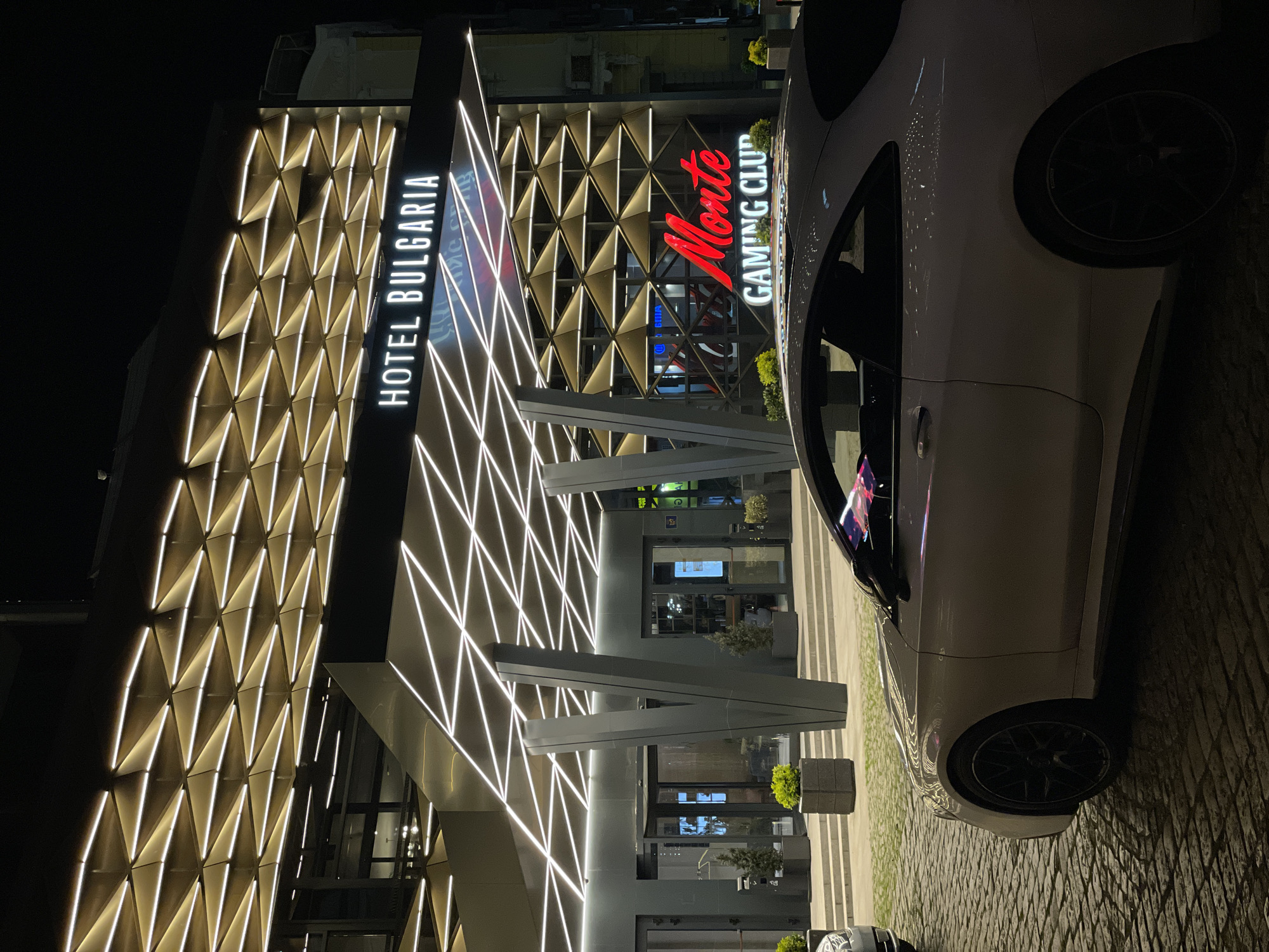 The image shows the exterior of Hotel Bulgaria in Burgas at night. The hotel's facade is illuminated, featuring a pattern of triangles with white lights. Adjacent to the hotel is the Monte Gaming Club, with its name in red neon lights. A white car is parked on the right side of the frame. The pavement around the car and the hotel entrance appears to be cobblestone. There are no visible people.