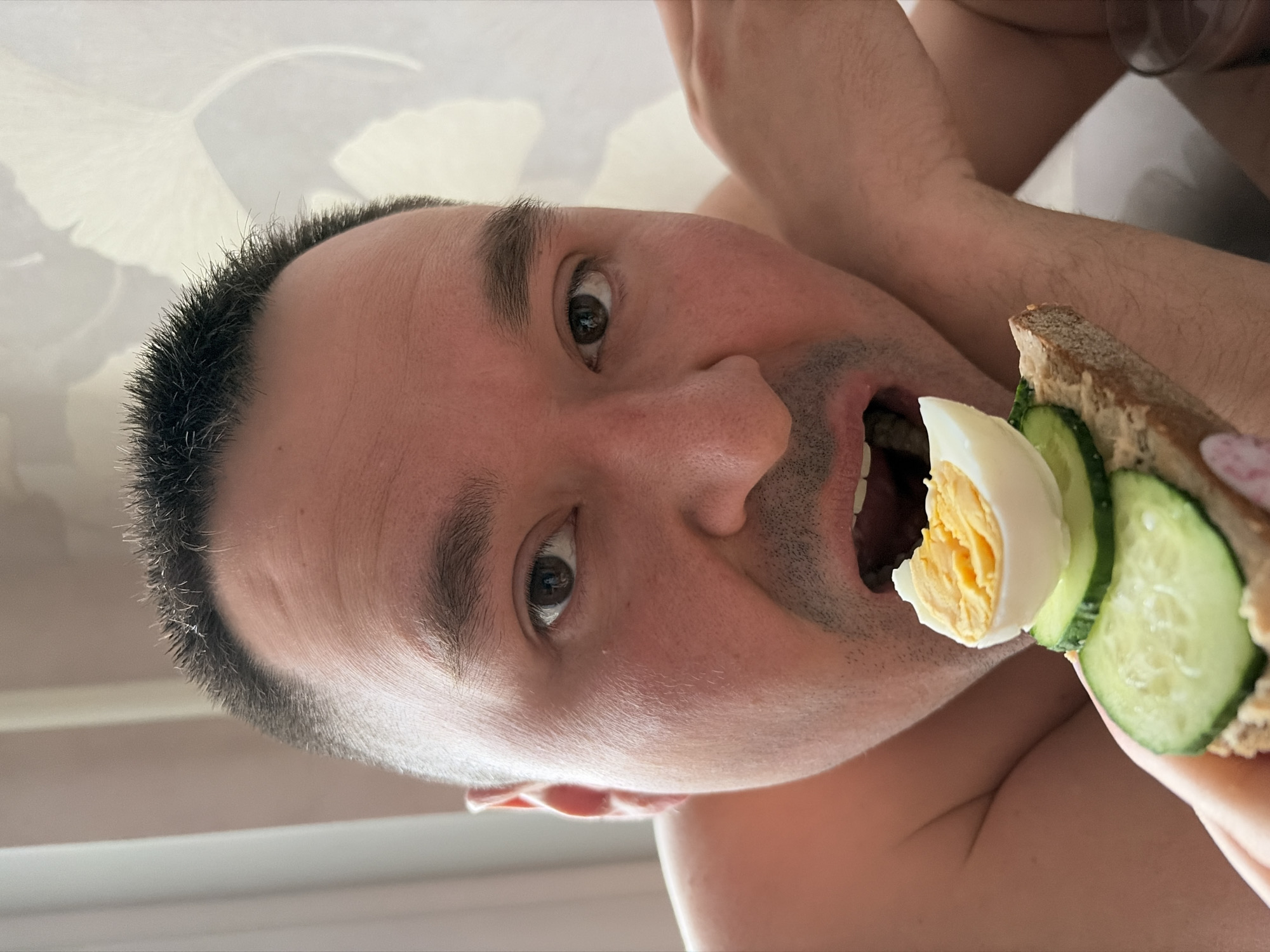 A close-up, tilted view shows a man eating a sandwich. The sandwich contains sliced hard-boiled egg and cucumber on brown bread. The man has dark hair, light skin, and is shirtless. The background is a light-colored wall. The location in the city of Izhevsk, Russia is not identifiable in the image.