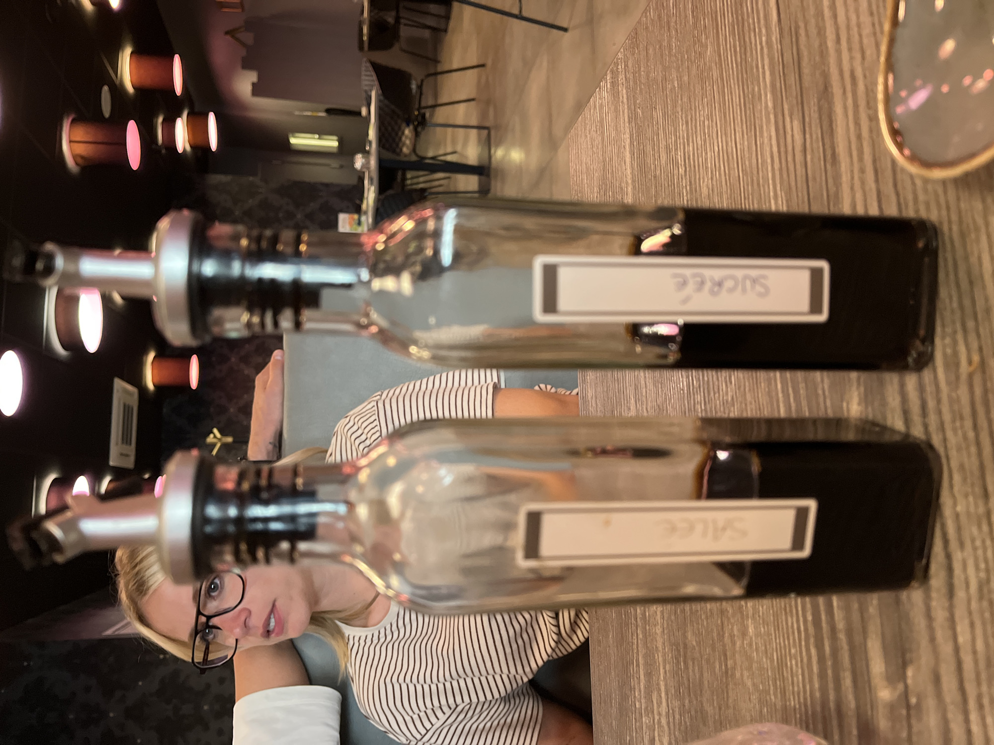 The image depicts a person sitting at a table with two bottles labeled "Salée" and "Sucrée". The bottles are likely filled with liquids like oil or vinegar. The person is wearing glasses and a striped shirt. The background suggests a restaurant or dining establishment. The bottles are placed on a wooden table. Based on the information available, the location is Andrézieux-Bouthéon, France.
