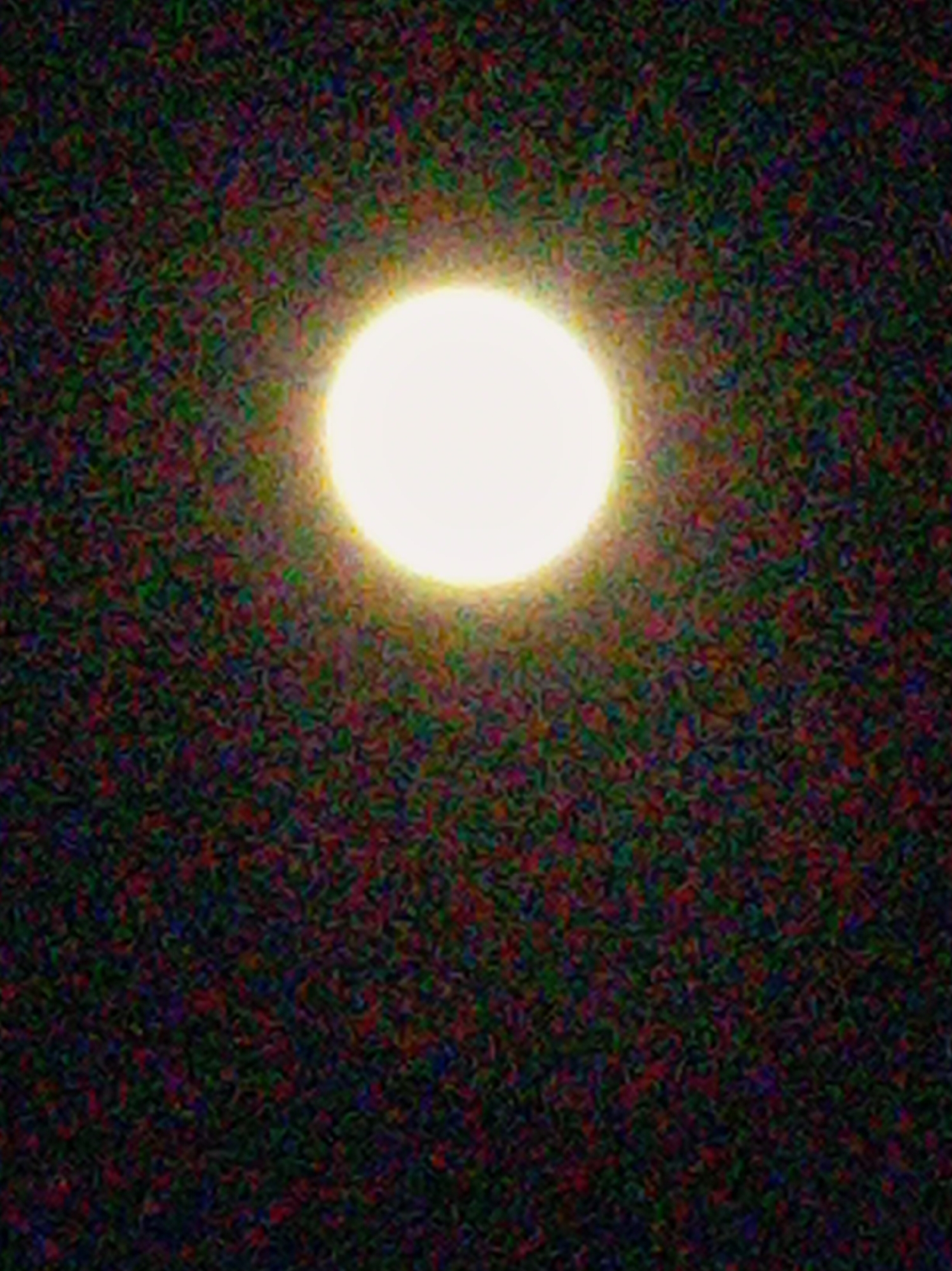 A photograph, possibly taken at night, shows a bright, circular light source in the center of the frame. The surrounding area is dark and appears speckled with a mixture of red, green, and blue pixels. The image is grainy and noisy. The subject is not related to the city of Artyom, Russia.