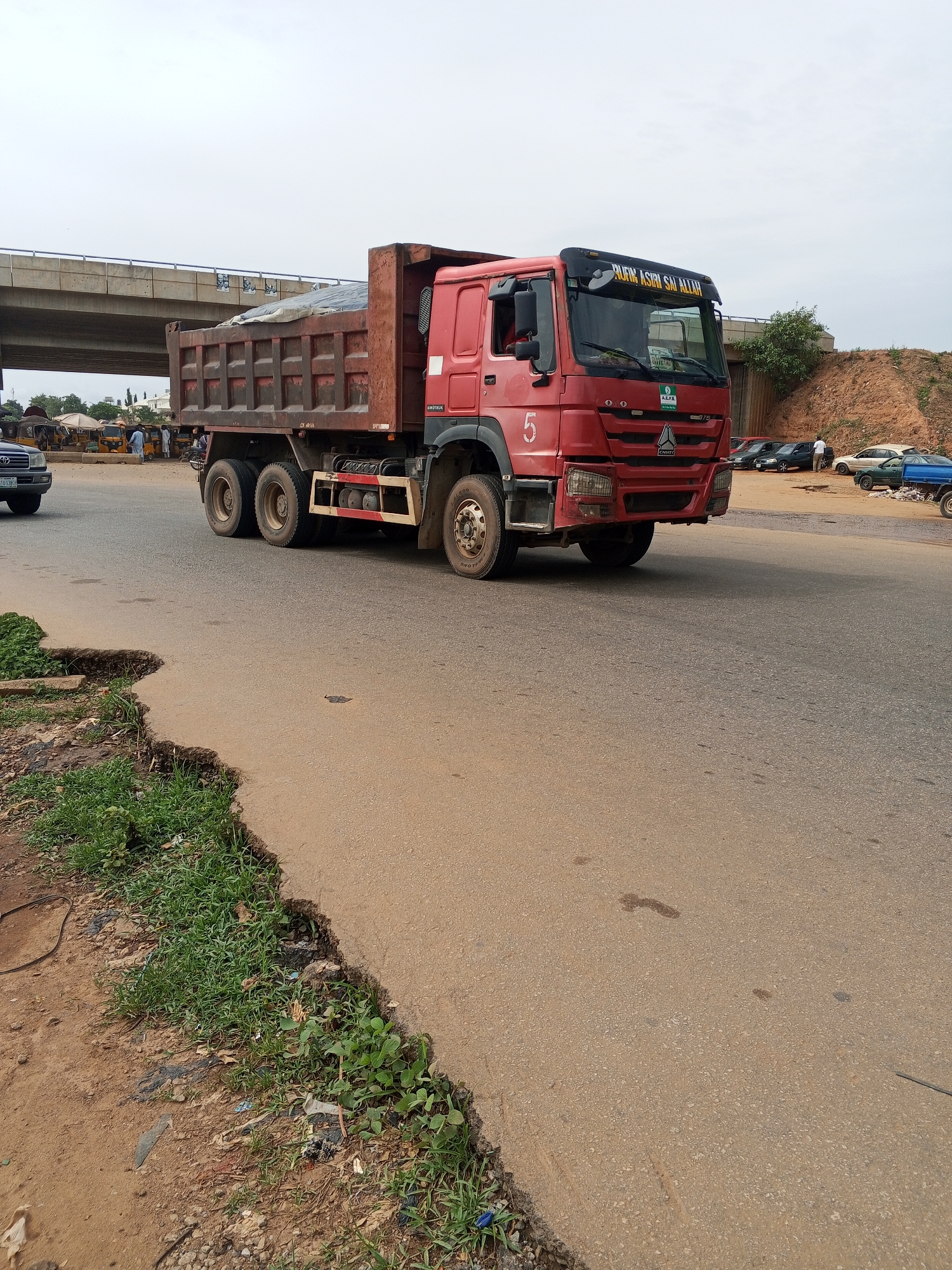 The image captures a red Sino Truck, a dump truck with the number "5" on the driver's side door, driving on a paved road in what appears to be Galadimawa Village, Nigeria. The road's edge shows signs of wear and disrepair with vegetation and debris. A bridge structure is visible in the background. In the distance, there's a gathering of people and vehicles, including tuk-tuks, suggesting a market or transportation hub. A number of parked cars also appear in the background on the right. A road appears to lead to the right of the truck. The sky is overcast.