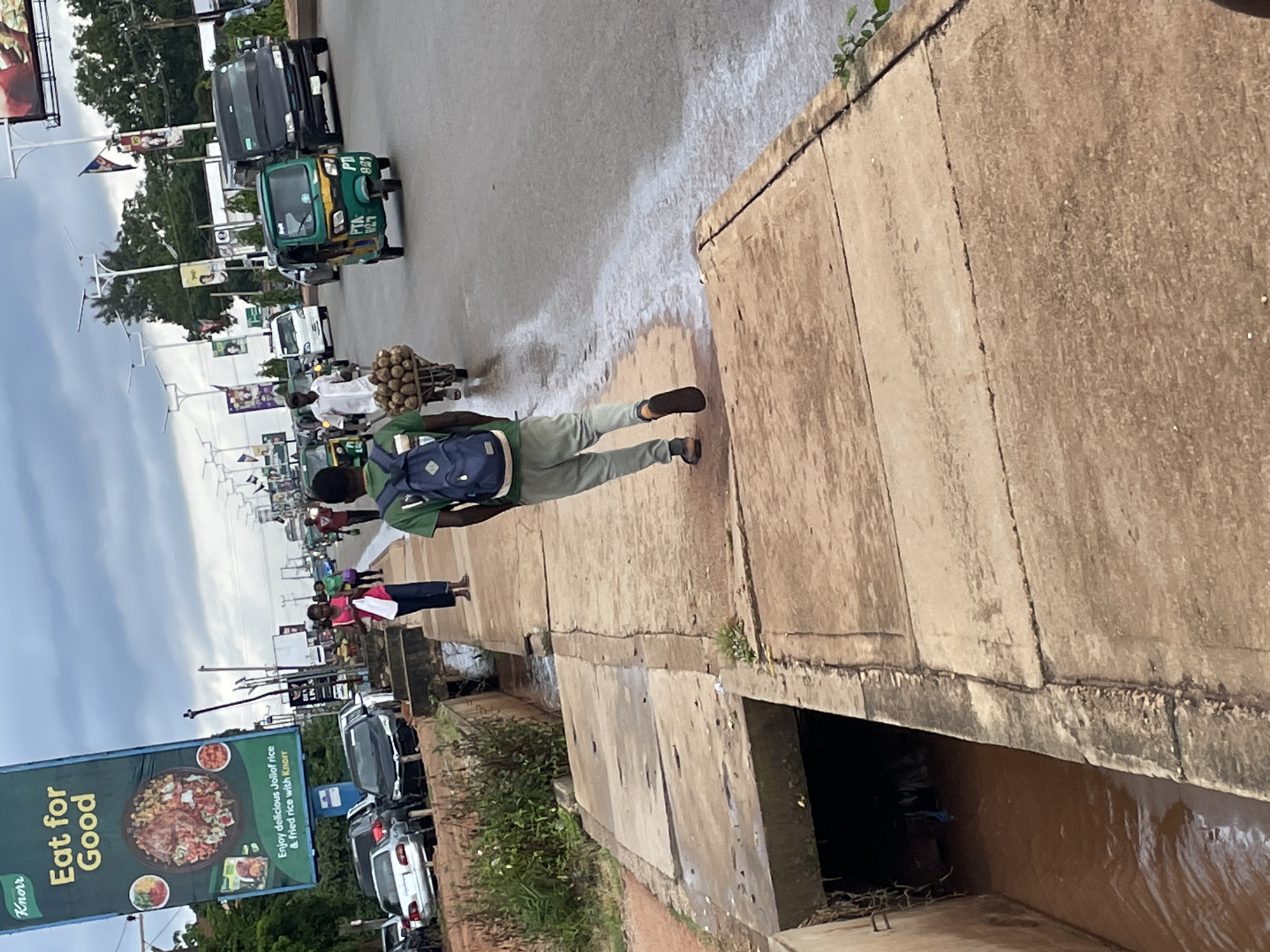 The image depicts a street scene in Jos, Nigeria, likely during the day with overcast skies. In the foreground, a person wearing a green shirt and gray pants, with a blue backpack, walks away from the camera on a concrete sidewalk. To the left of the sidewalk is a concrete drainage channel with murky, reddish-brown water flowing through it. A large billboard for Knorr, promoting "Eat for Good" and Jollof rice, is visible on the left side of the image.
The street itself is paved and shows signs of wear. Several vehicles are present on the road, including cars, a black SUV, a green three-wheeled auto-rickshaw (Keke Napep), and other commercial vehicles. Pedestrians are seen on the sidewalk and in the street. A person is pushing a cart laden with what appears to be produce, possibly yams, on the right side of the road. Traffic lights and streetlights are visible, indicating an urban environment. The overall impression is one of a busy, everyday street scene in Nigeria.