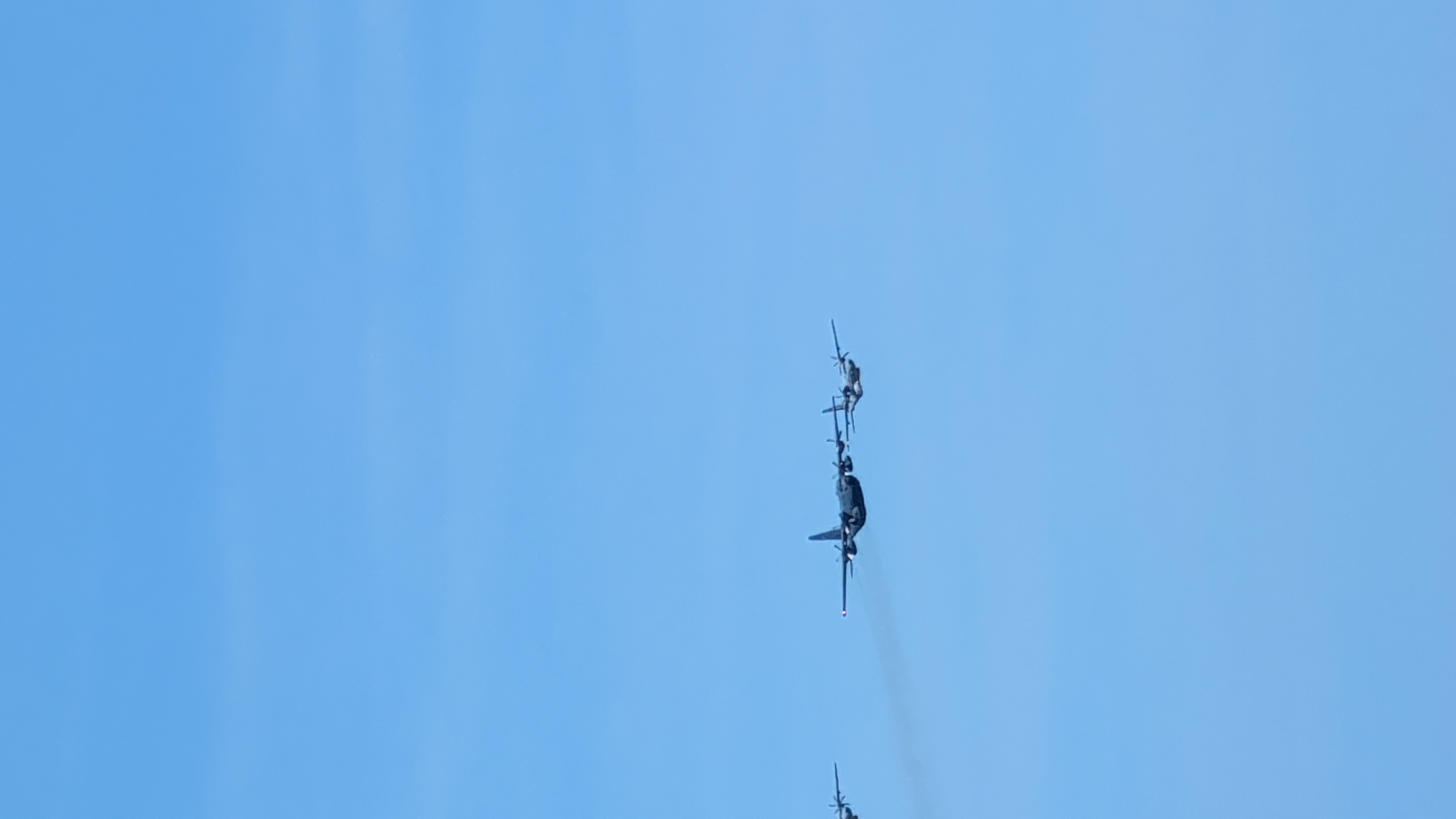 An aerial view captures a formation of four transport aircraft flying against a clear, pale blue sky. The planes are positioned at varying distances and heights, with three of them in a tighter cluster. The lead aircraft in the cluster is dark and appears to have four engines. Behind it, another similar aircraft is visible, followed by a smaller, propeller-driven aircraft. To the far left, a fourth aircraft, also dark and appearing to be a transport plane, flies at a lower altitude and further away from the main group. No ground features or city identification are visible in this image, which is solely focused on the aircraft in flight against the sky. The image does not depict any specific location in Warsaw, Poland, or any other identifiable geographical area.