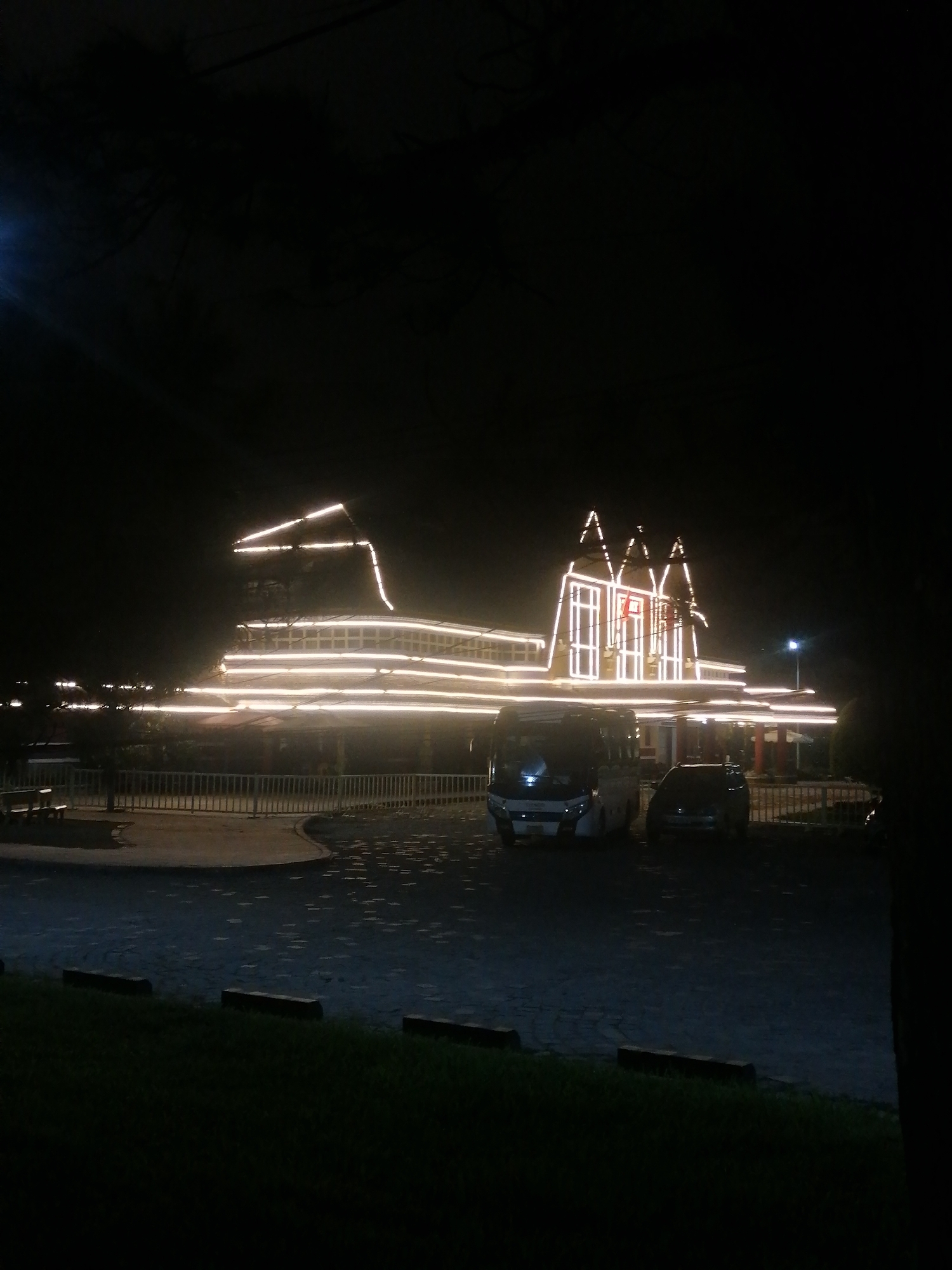 This is a nighttime photograph of the entrance to the Dalat Palace Hotel in Phường Xuân Hương - Đà Lạt, Vietnam. The building is illuminated with numerous string lights, highlighting its architectural features, including triangular roof elements and horizontal banding. In the foreground, a paved parking area is visible, with two vehicles parked: a white van on the left and a dark-colored SUV on the right. Beyond the parking area, a metal fence separates the hotel grounds from a darker, grassy area. Trees are present on the left side of the image, partially obscuring the hotel's facade and the night sky. The overall impression is one of a well-lit, possibly festive, establishment at night.