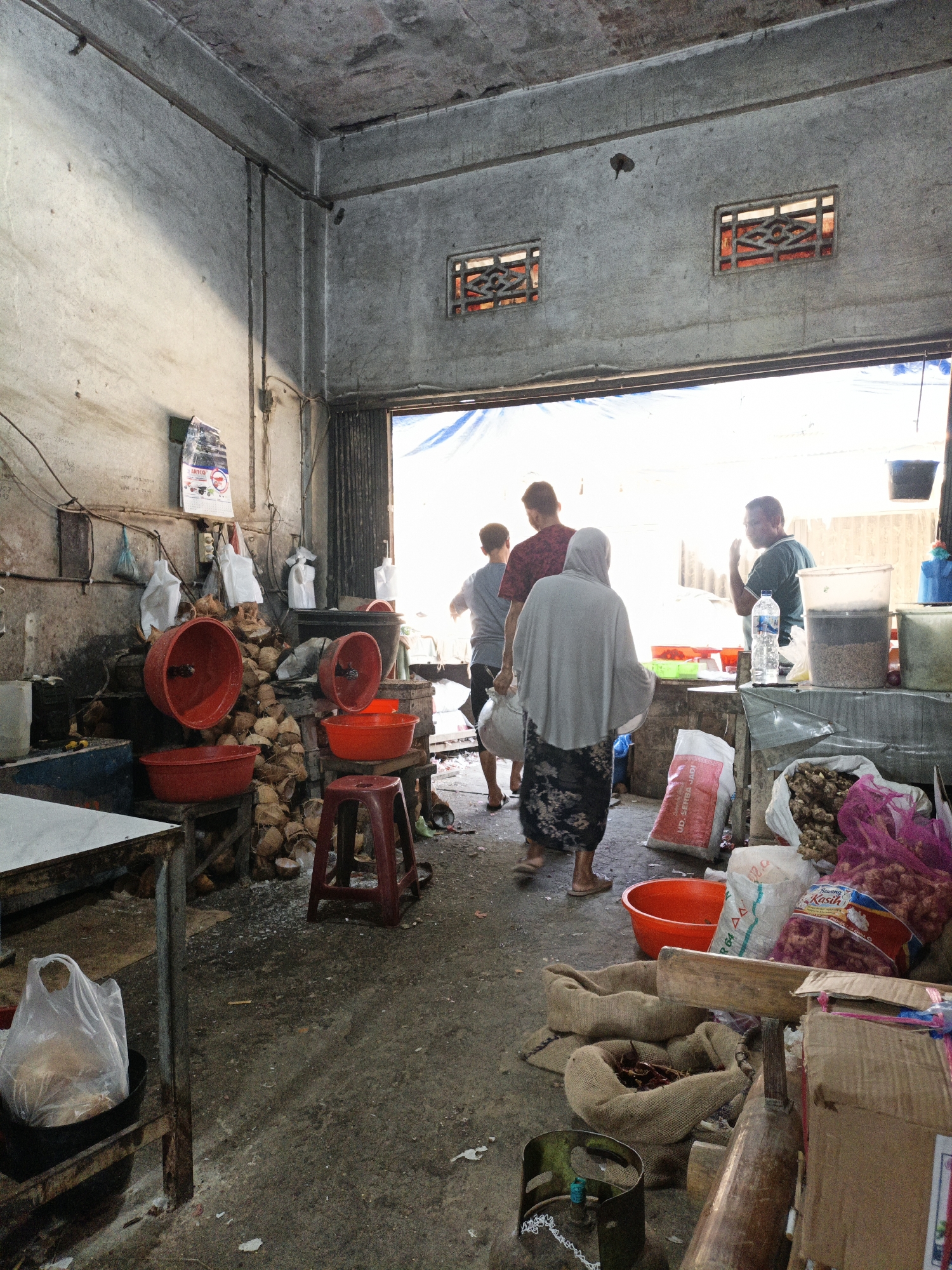 This is a market stall in Keudegeudong, Indonesia, with a concrete floor and rough concrete walls. The space appears open-air at the back, with a blue tarp providing overhead cover. Several people are present, engaged in market activities.
In the foreground, a metal table with a white top is on the left, with a plastic bag containing coconuts placed in a black basin beneath it. To the right of this table, a red plastic stool is visible. Behind the stool, a pile of coconuts and their husks is situated against the wall, with several large red basins stacked or placed nearby. Some of these basins appear to be empty, while others may contain items. A calendar is hanging on the wall above the coconuts.
Further into the stall, a group of individuals are visible. One person, wearing a grey hijab and a patterned skirt, walks away from the camera towards the open back of the stall. Another person, in a dark shirt and possibly carrying something, walks in the same direction. A third person, wearing a red and black patterned shirt, is also moving away from the camera.
In the background, a man in a green shirt is standing and appears to be talking or gesturing. Next to him is a clear plastic container filled with a dark substance and a bottle of water on a table. Various sacks and bags of produce are arranged around the stall, including burlap sacks filled with what appear to be spices or dried goods, and pink mesh bags containing red onions or similar items. Cardboard boxes are also stacked in the vicinity. The overall impression is of a bustling, functional marketplace with goods laid out for sale.
